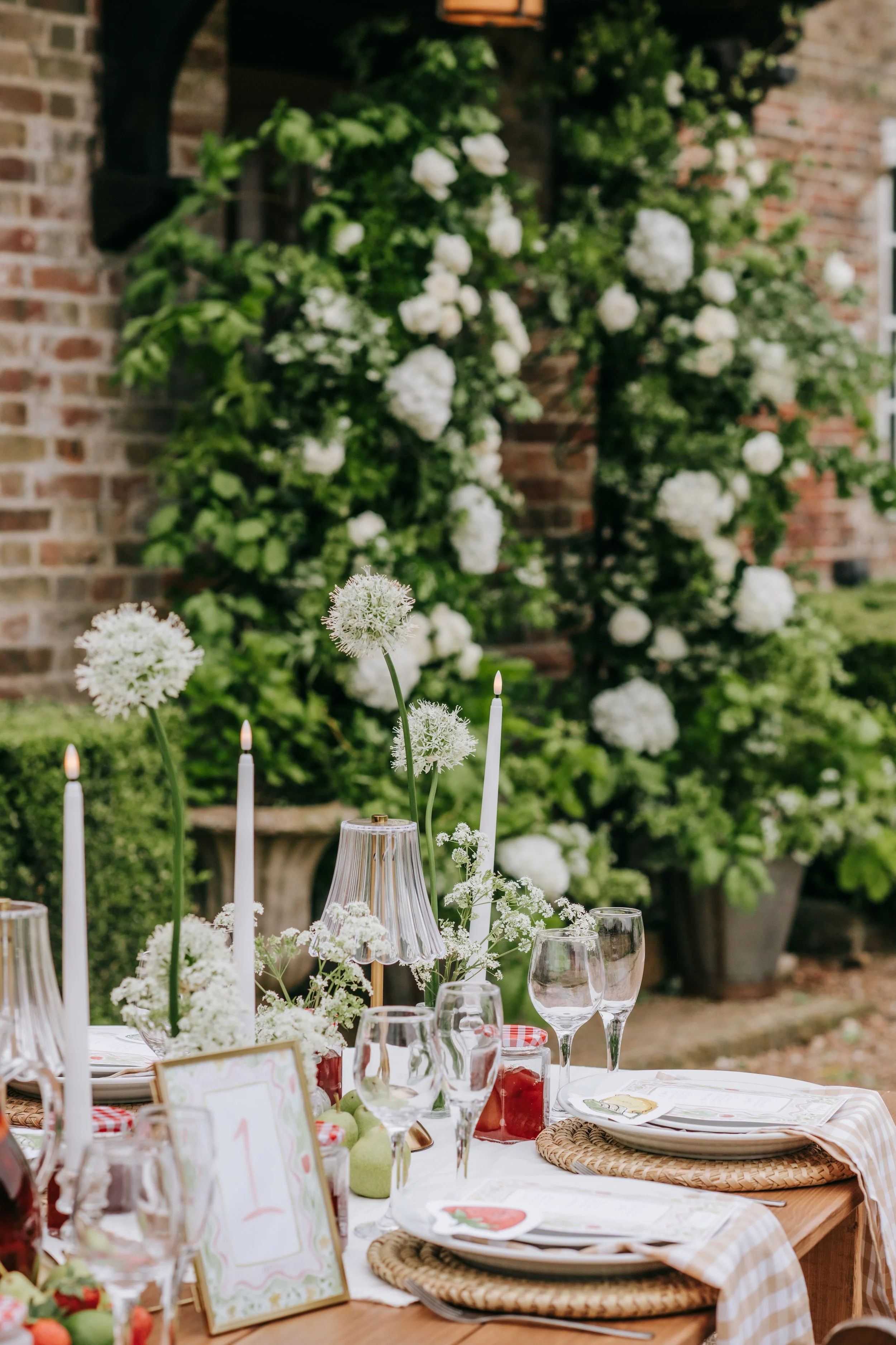 Outdoor table set for a celebration with white floral centerpiece, candles, glassware, and place settings, with greenery and flowers in the background.