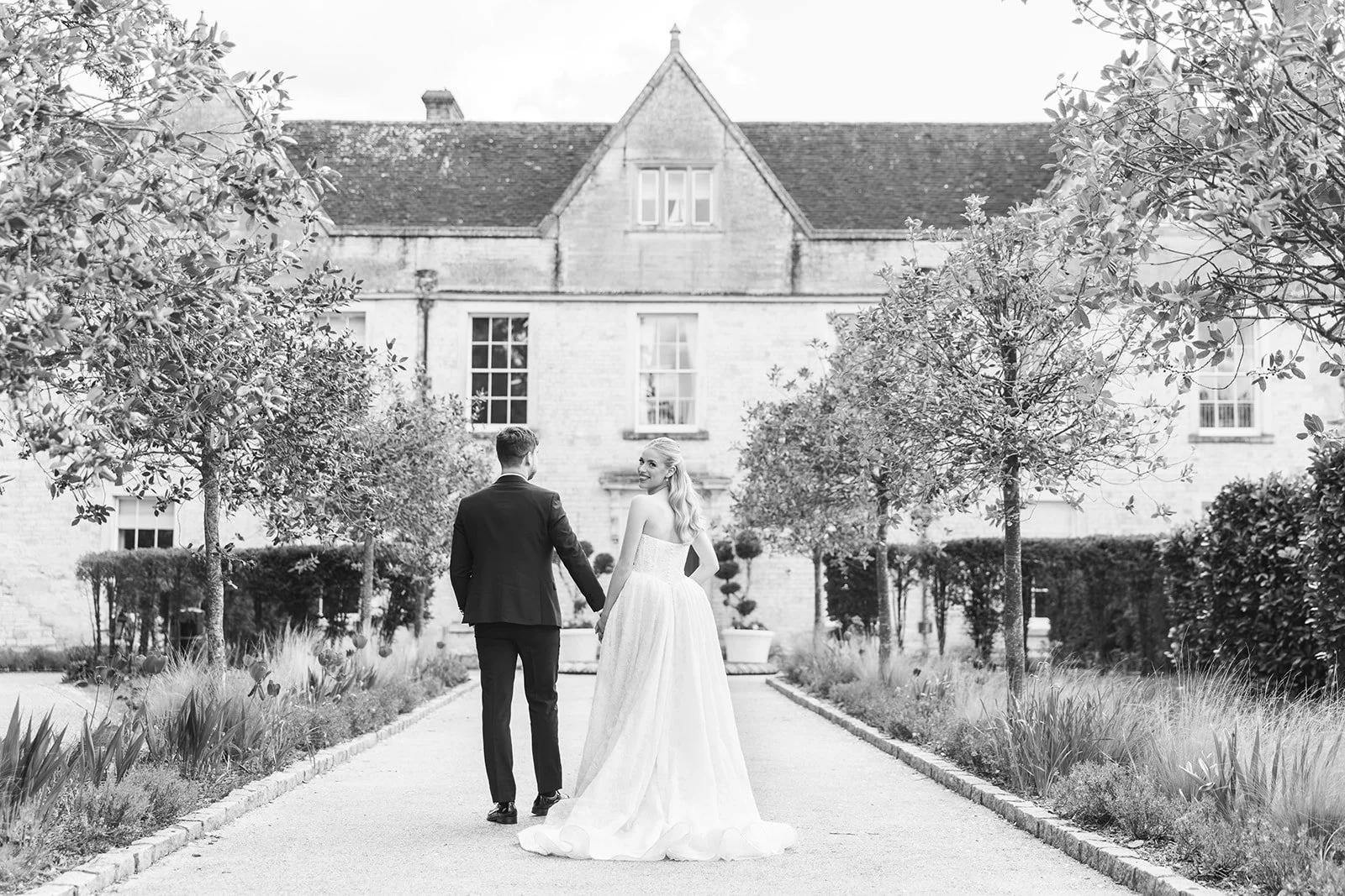 Black and white photo of a bride and groom walking hand in hand along a garden path, with a large house in the background.
