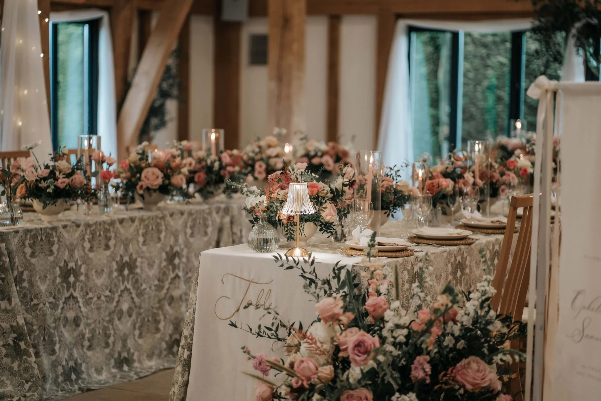 A beautifully decorated wedding reception table with pink and white floral arrangements, candles, and an elegant lace tablecloth inside a rustic venue with exposed wooden beams and large windows.