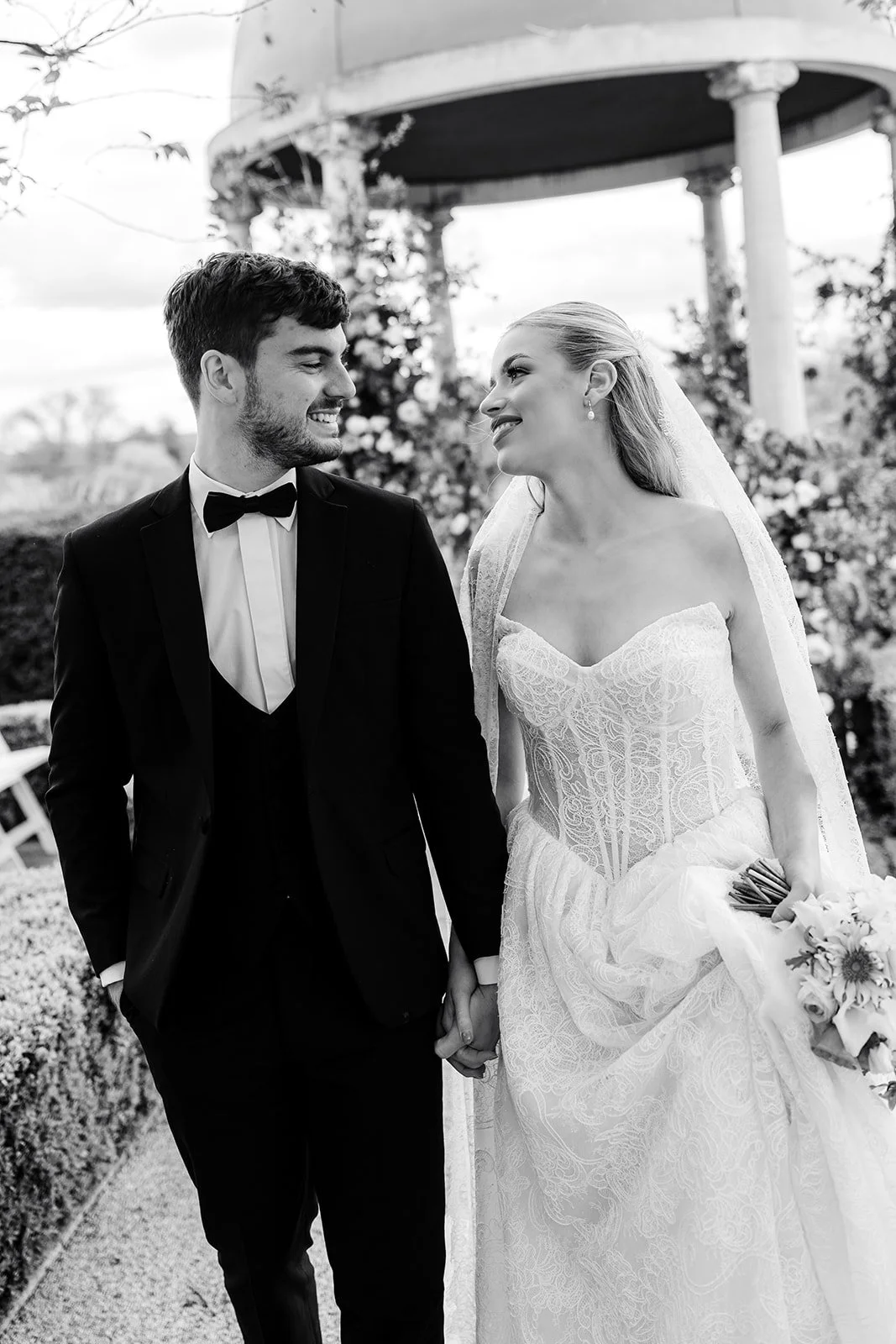 A black and white photograph of a bride and groom holding hands, smiling, and looking at each other outdoors during their wedding.