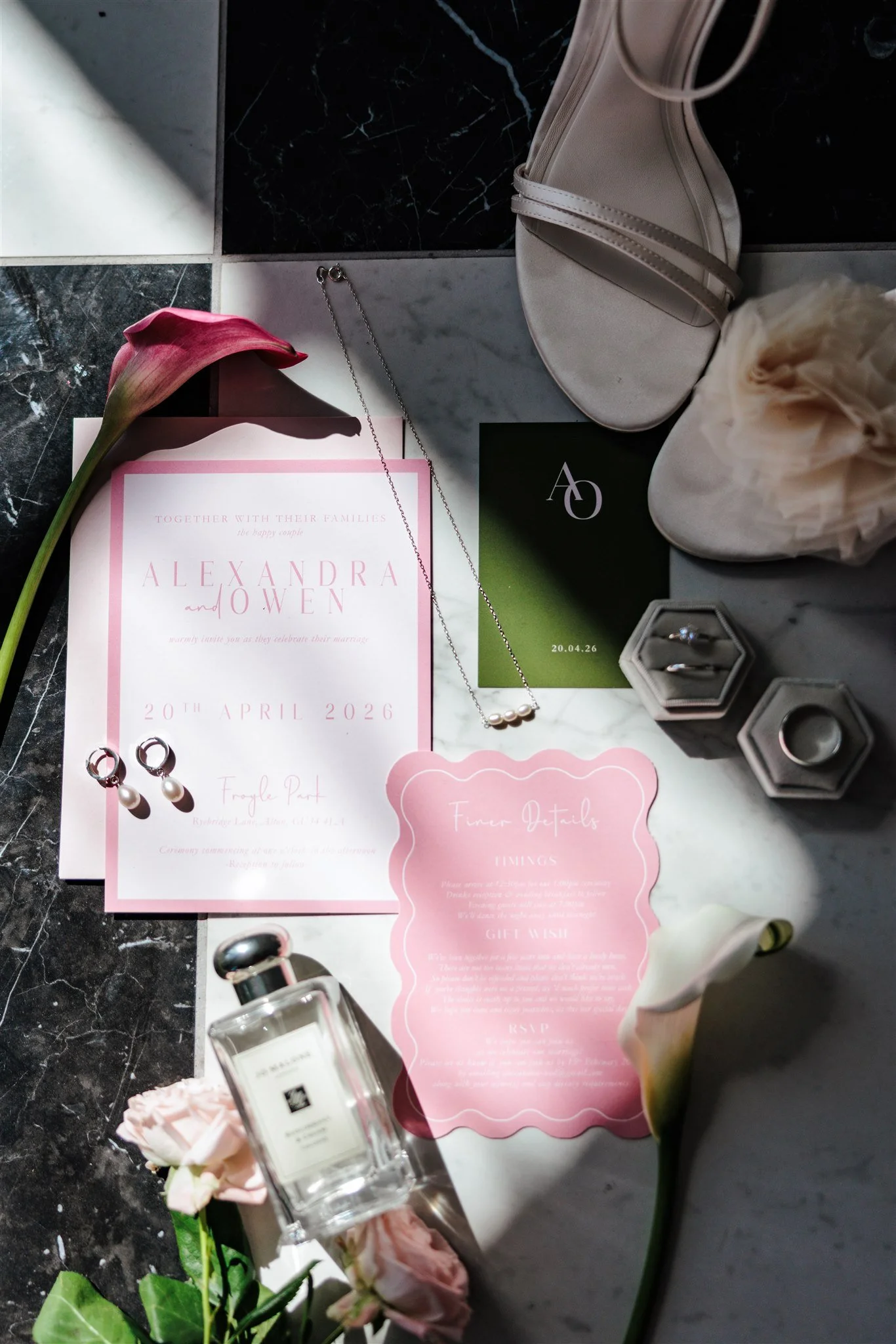 Wedding invitation card for Alexandra and Owen dated April 20, 2026, surrounded by pink flowers, jewelry, a white purse, and a perfume bottle on a marble and black tiled surface.