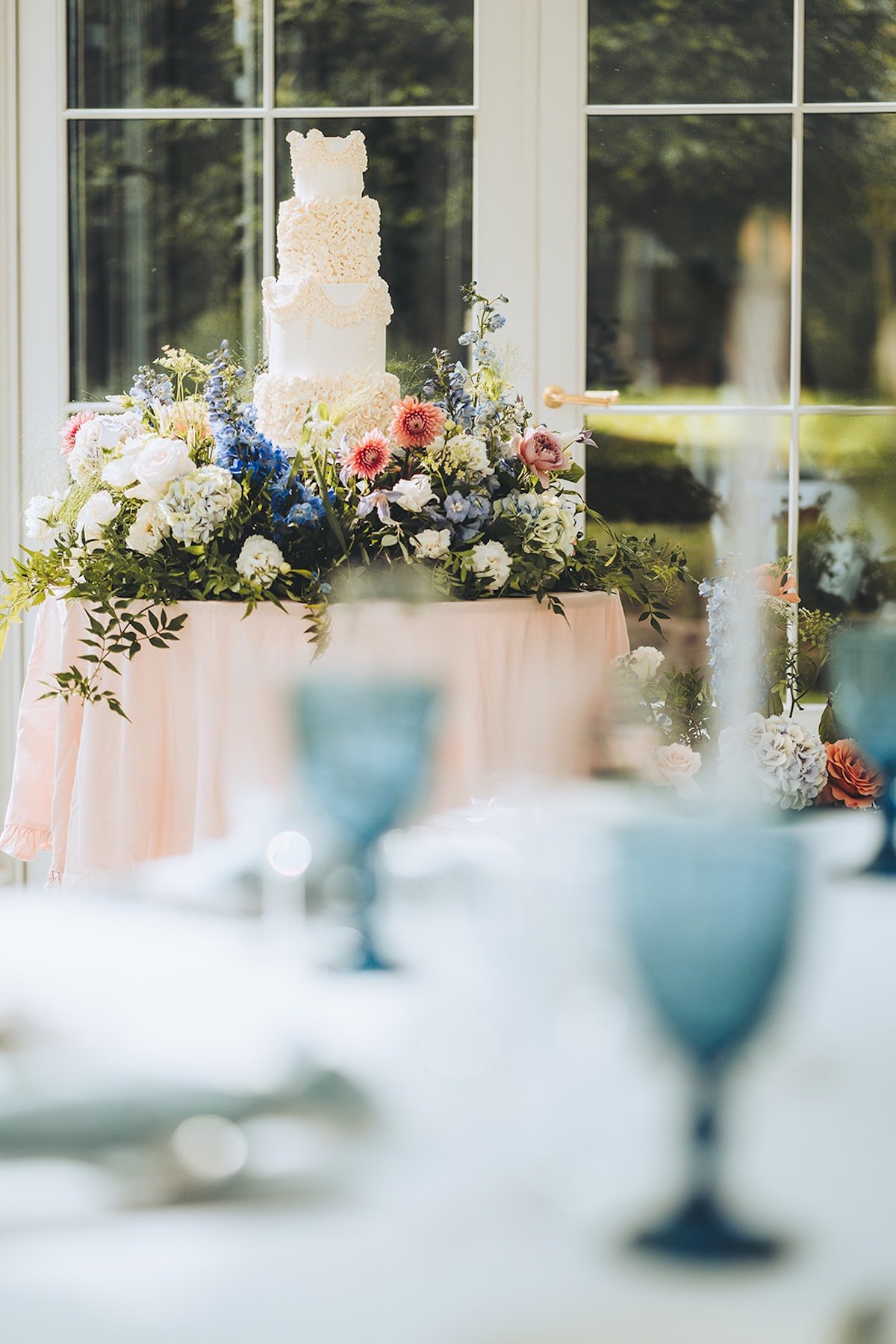 A wedding cake with cascading floral decorations on a table with pink and white fabric, surrounded by a variety of flowers, placed near a window with a view of greenery outside.