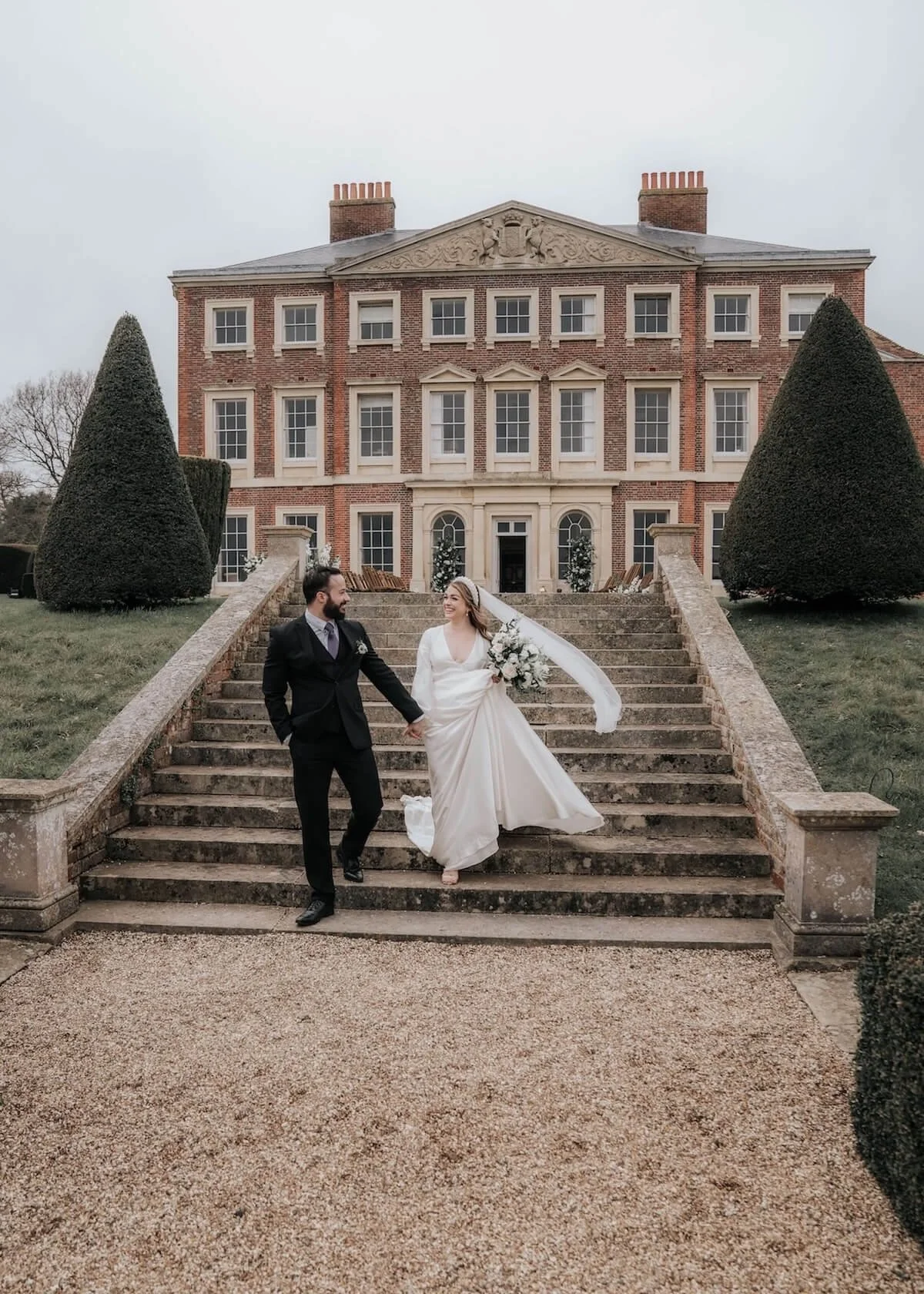 A bride and groom walking down the steps in front of a large, historic brick mansion with manicured trees and stone steps, holding hands and smiling.