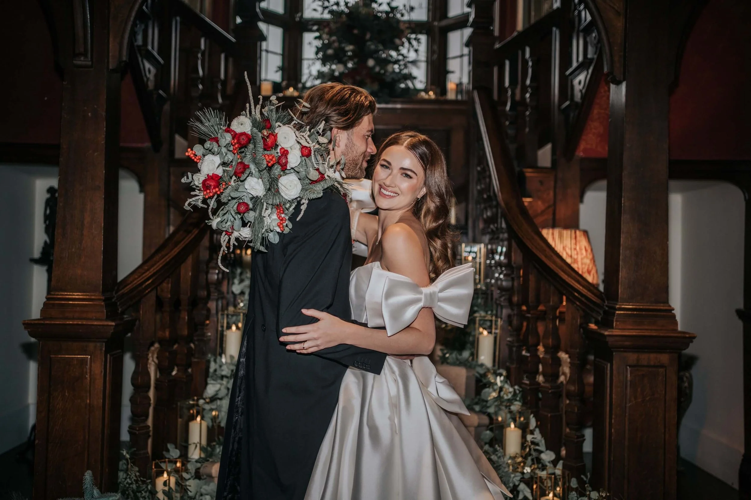 A newly married couple embracing in front of a decorated staircase with candles and floral arrangements, Christmas tree in the background, in a historic wooden venue.