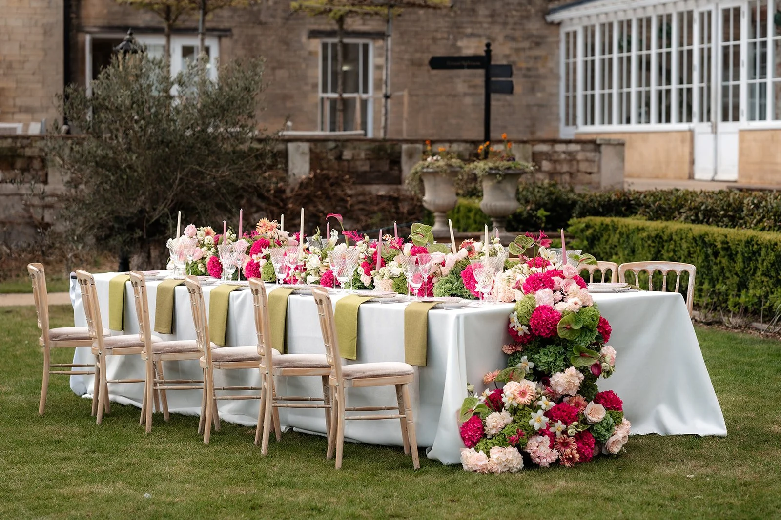 A long rectangular outdoor dining table decorated with pink, white, and green floral arrangements, set with glassware, white plates, and pink candles in glass holders, on a lawn with a stone building in the background.