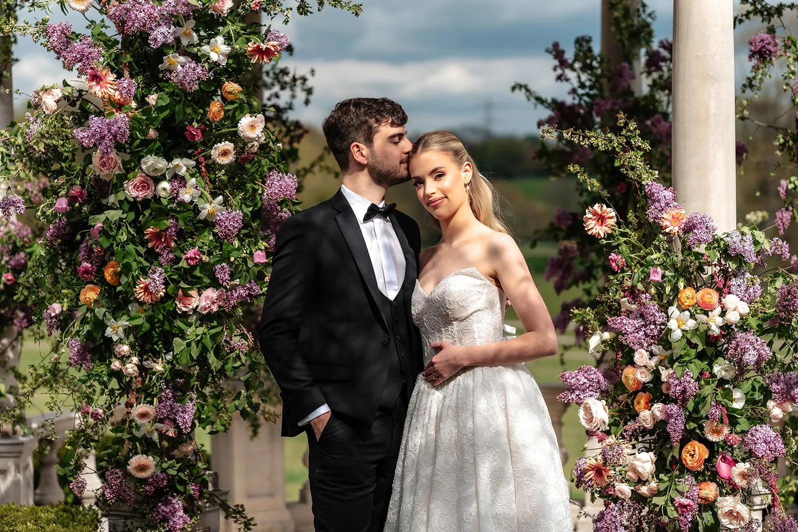 A newlywed couple posing beneath a floral arch decoration outdoors. The groom in a black tuxedo, and the bride in a white wedding dress, are standing close together, with the groom whispering into the bride's ear as she smiles at the camera.