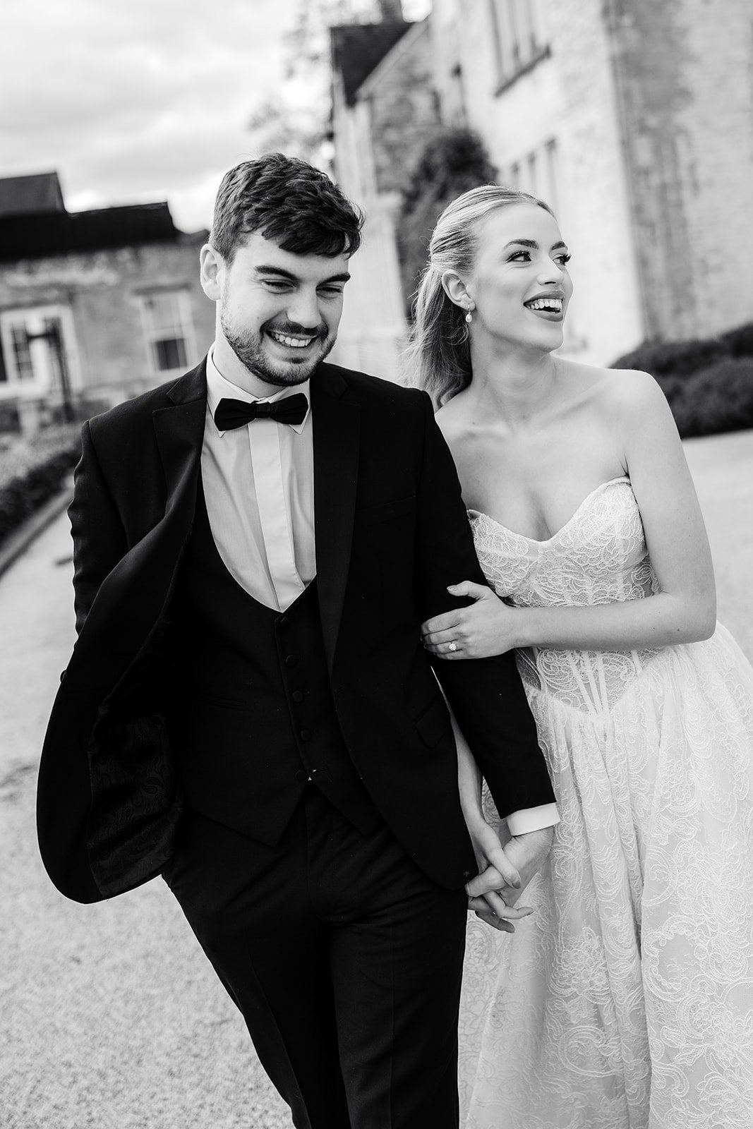 A black and white photo of a happily married couple walking outdoors, holding hands. The man is wearing a tuxedo with a bow tie, and the woman is in a strapless lace wedding dress. They are smiling and looking joyful.