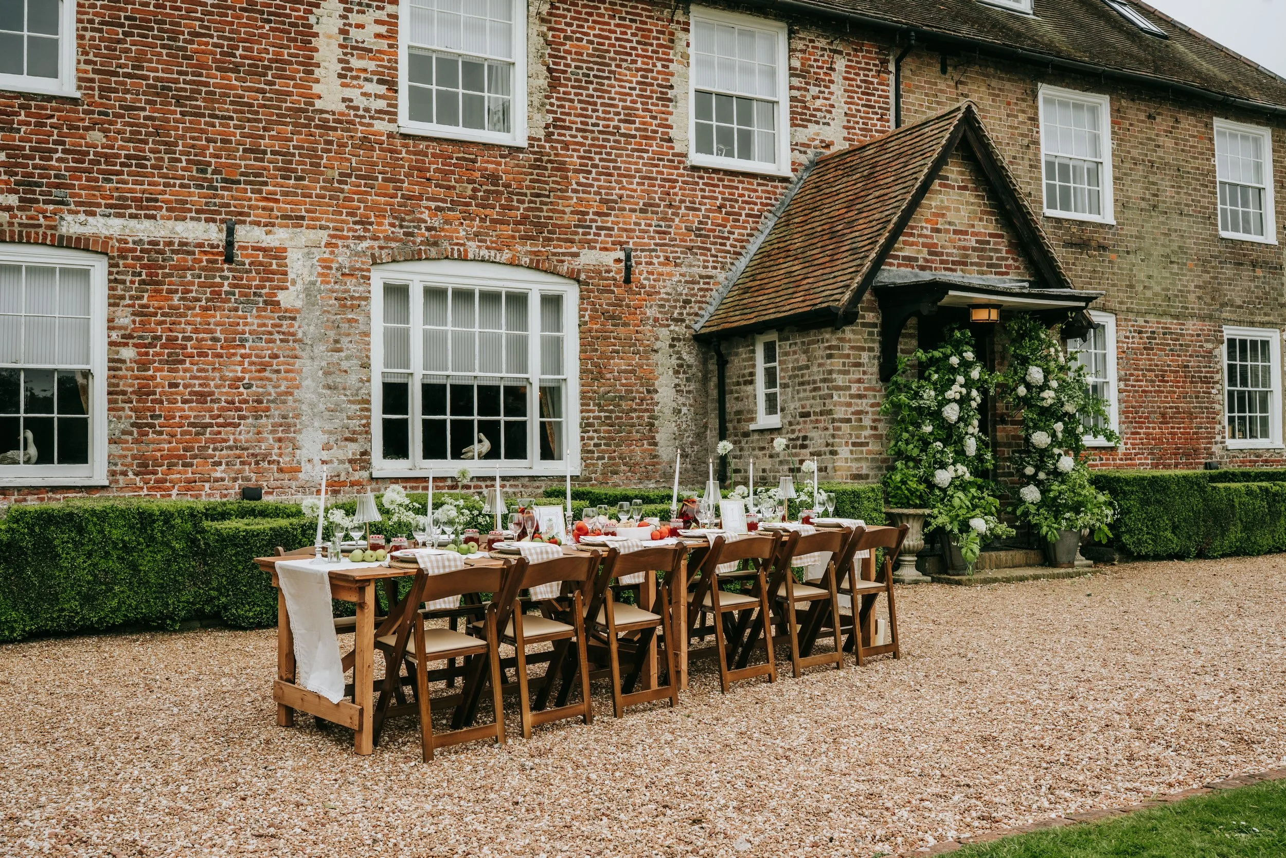 Outdoor dining setup in front of a brick house, with a long table, wooden chairs, tablecloth, candles, and autumn-themed decorations, with a hedge and white flowers nearby.
