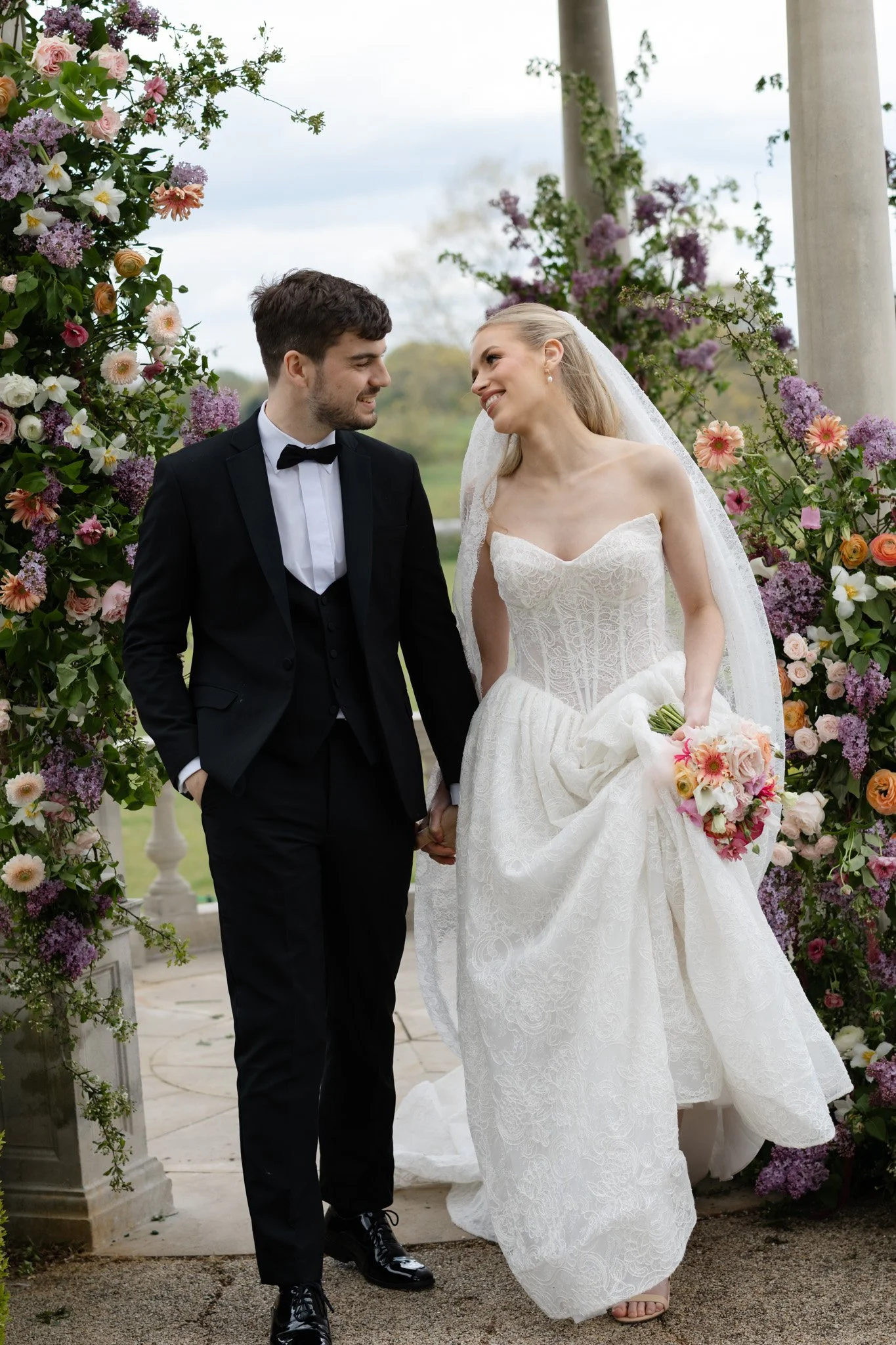 A bride and groom holding hands and smiling at each other under a floral arch at their outdoor wedding ceremony.