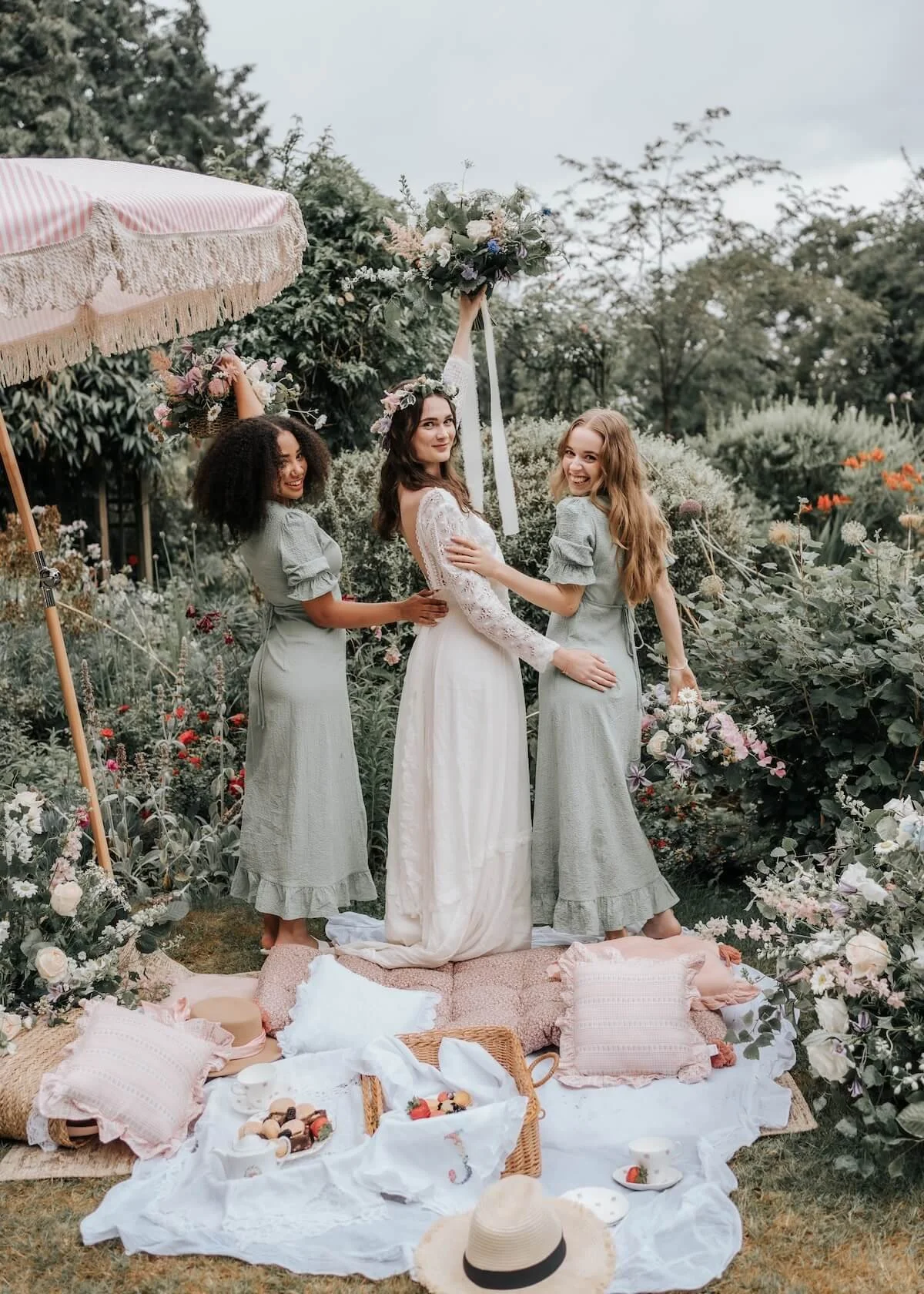 Three women at a garden wedding, with one woman in a white bridal dress holding a floral bouquet high, while the other two women in pastel dresses stand beside her, all smiling happily amidst lush greenery and flowers. There is a picnic setup with pi