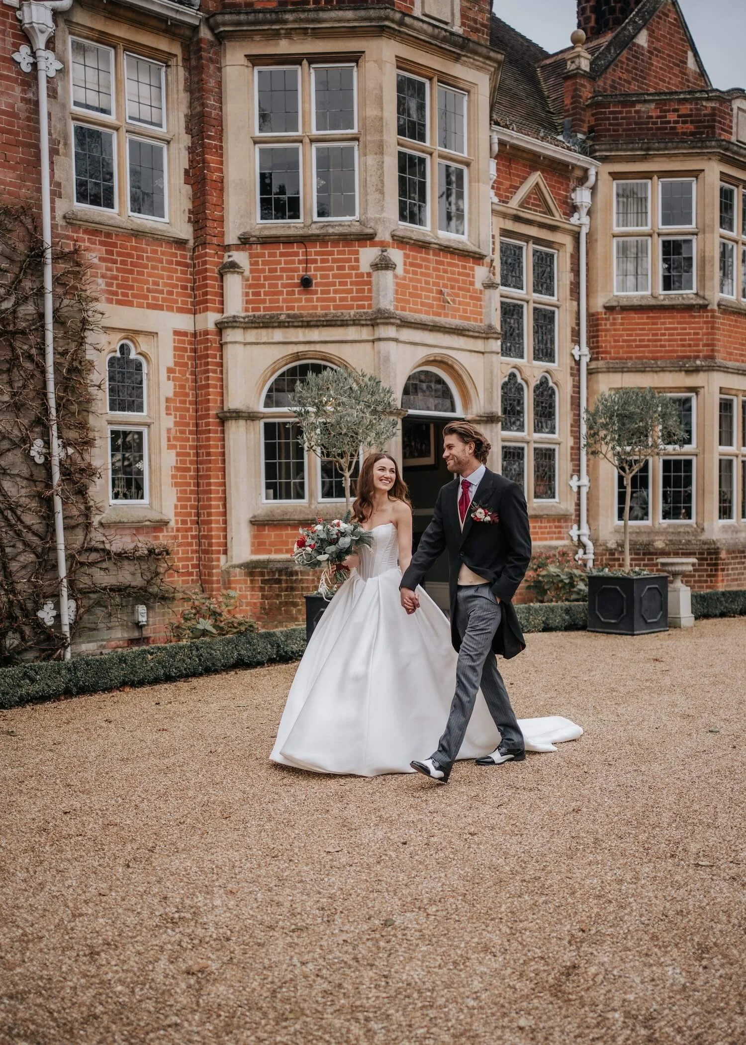 A bride and groom holding hands and walking outside a large brick and stone historic building with multiple bay windows and a sloped roof, during daytime.