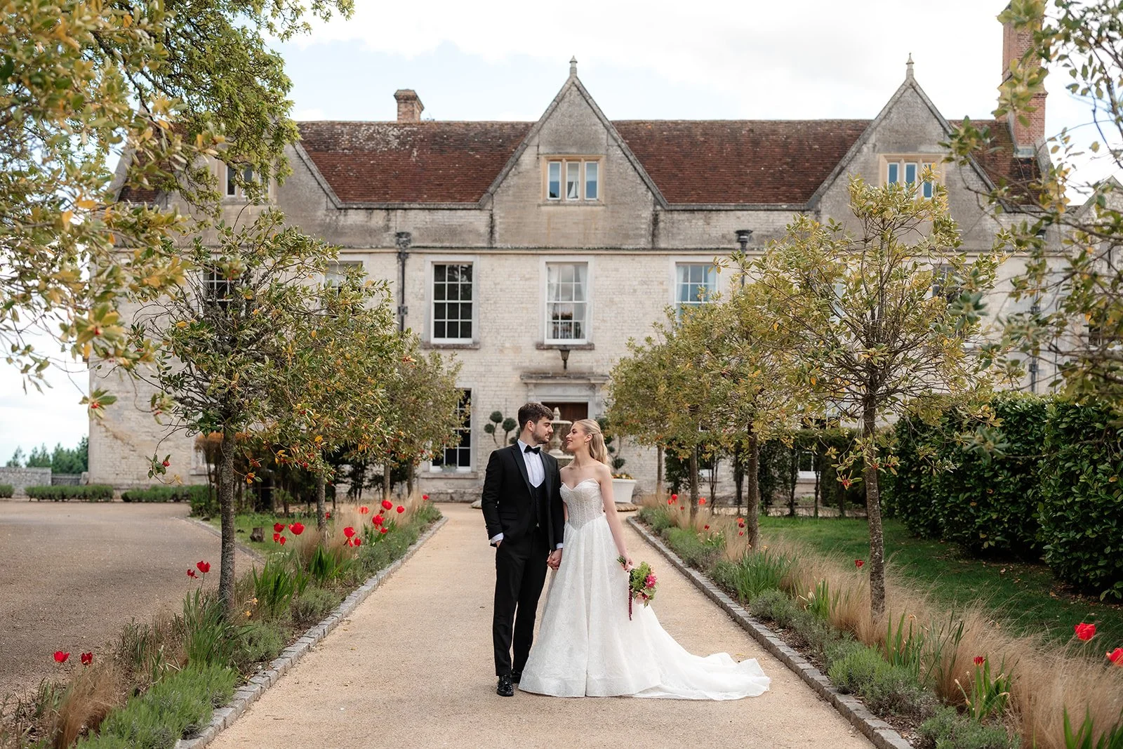 A bride and groom holding hands, standing on a garden pathway in front of a historic stone mansion. The bride is in a white wedding gown holding a bouquet, and the groom is in a black tuxedo. The garden features trees, red flowers, and landscaped greenery.