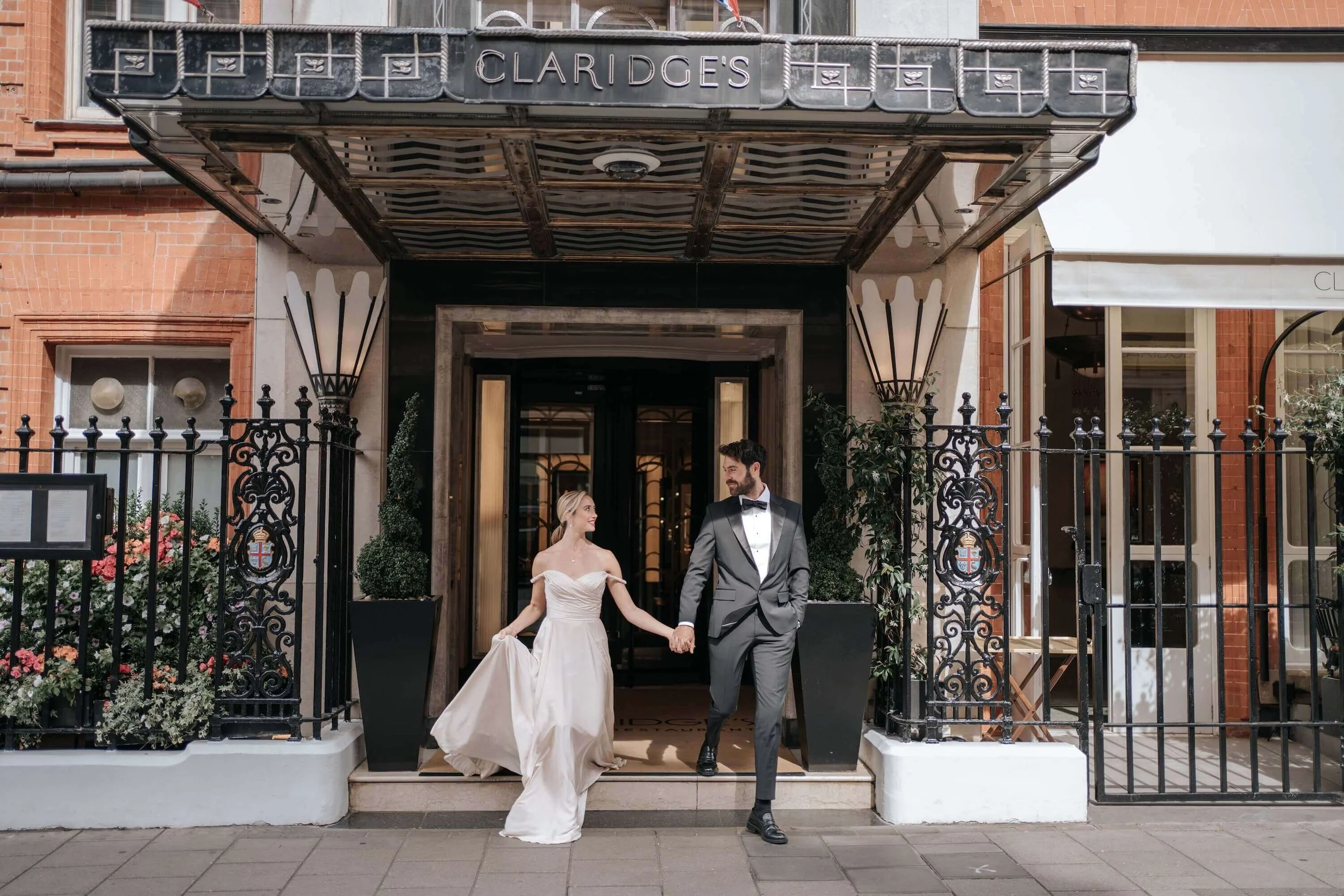 A bride and groom in wedding attire holding hands and walking out of a building labeled 'Claridge's'.
