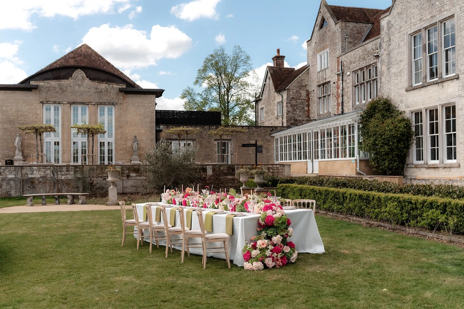 Outdoor wedding reception setup with a long table decorated with pink and white flowers, surrounded by chairs, on a well-manicured lawn in front of an historic stone building with large windows and a fence with potted statues.