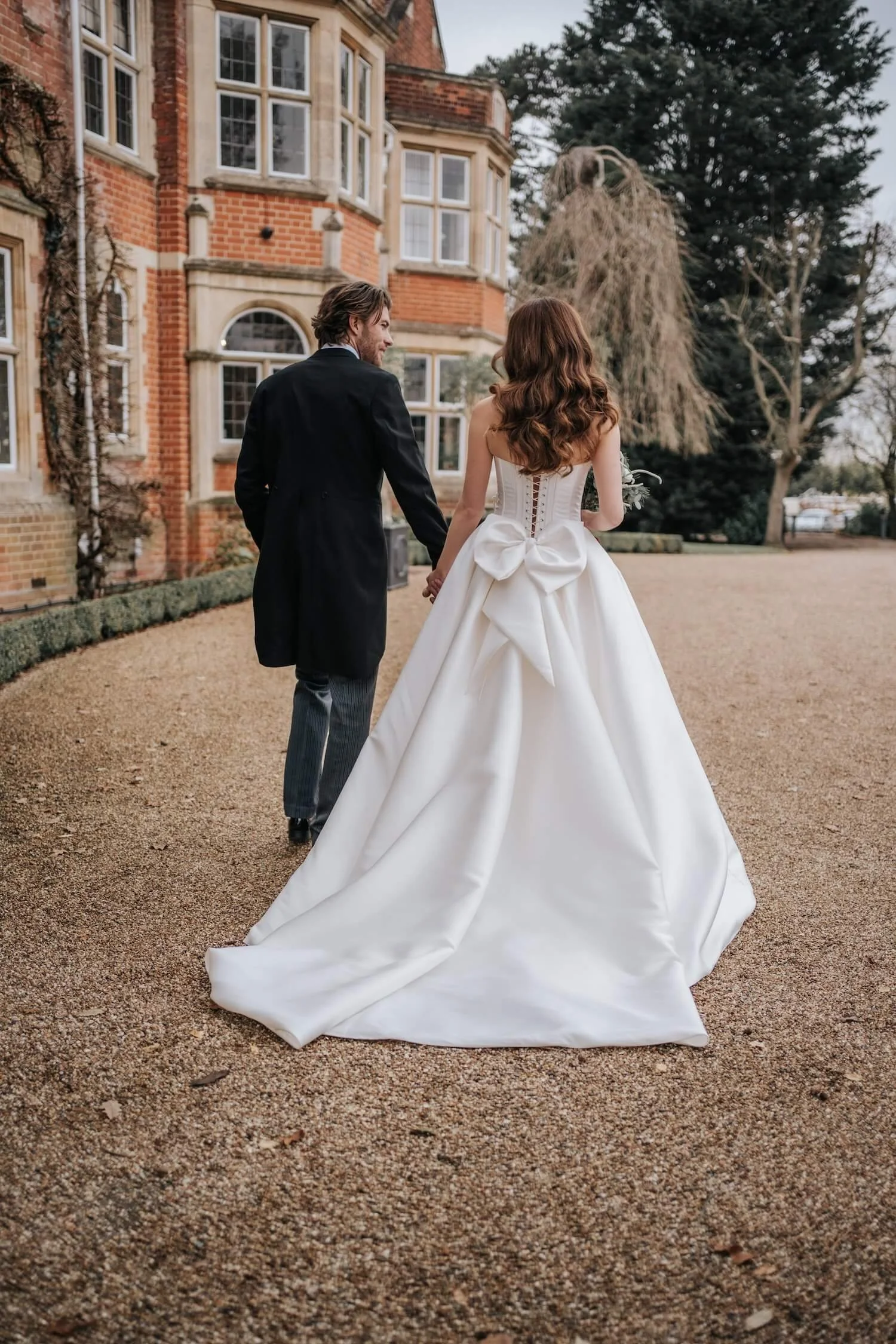 A bride and groom holding hands, walking away from the camera. The bride is wearing a white wedding gown with a large bow at the back, and the groom is dressed in a black suit. They are outdoors in front of a large brick building with multiple windows, and there are trees around them.