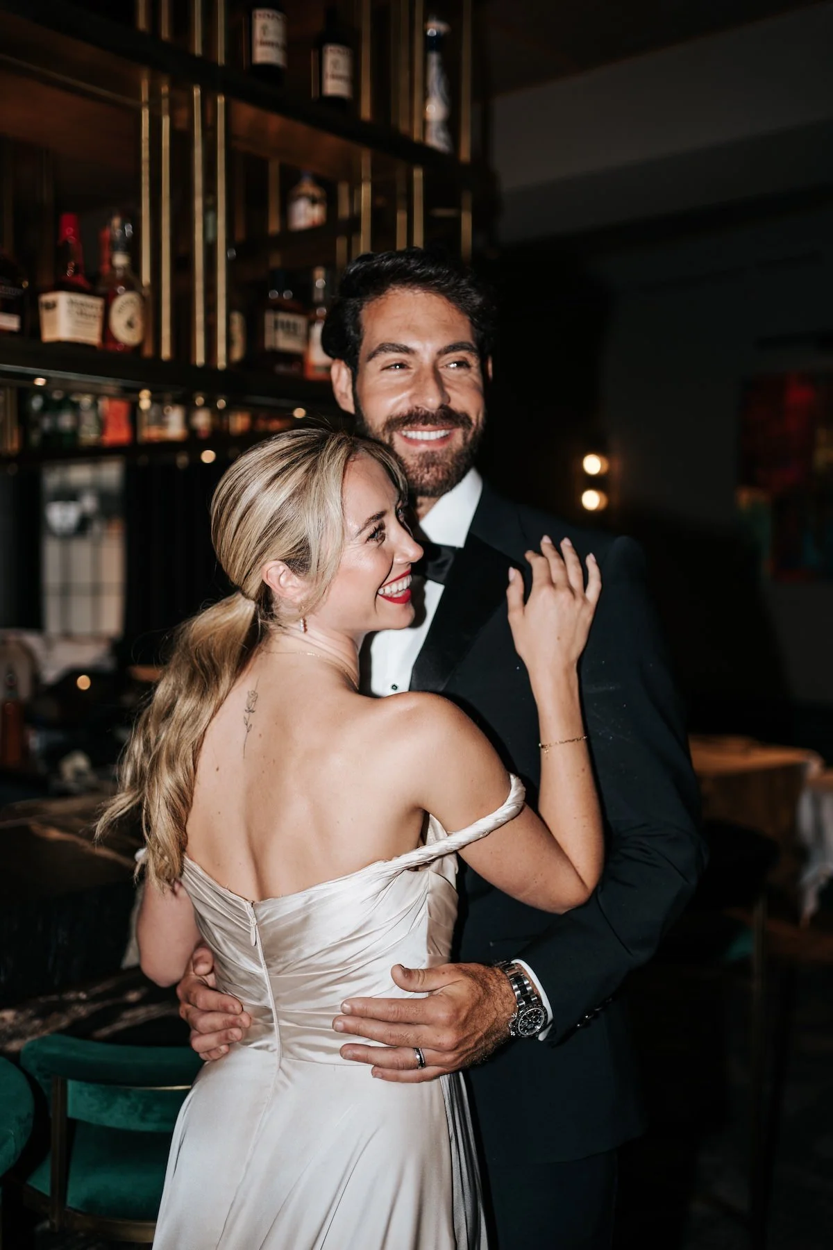 A couple dressed in formal attire dancing closely and smiling at each other in a dimly lit bar or restaurant with wood shelves and liquor bottles in the background.