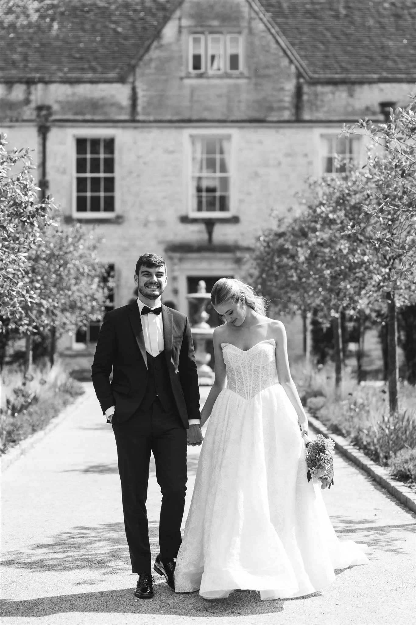 A smiling man in a tuxedo and a woman in a wedding dress walking hand in hand on a pathway surrounded by trees and a large house in the background.
