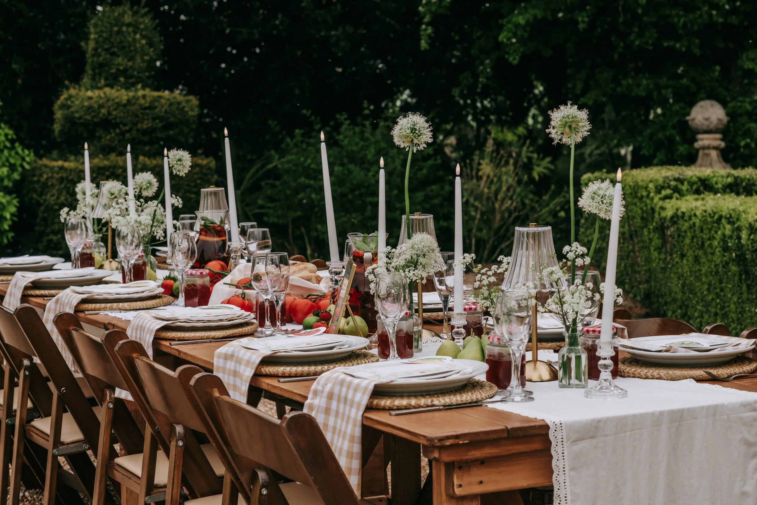 Outdoor dining table set with plates, glasses, white candles, flowers, and fruits, surrounded by wooden chairs on a garden patio.