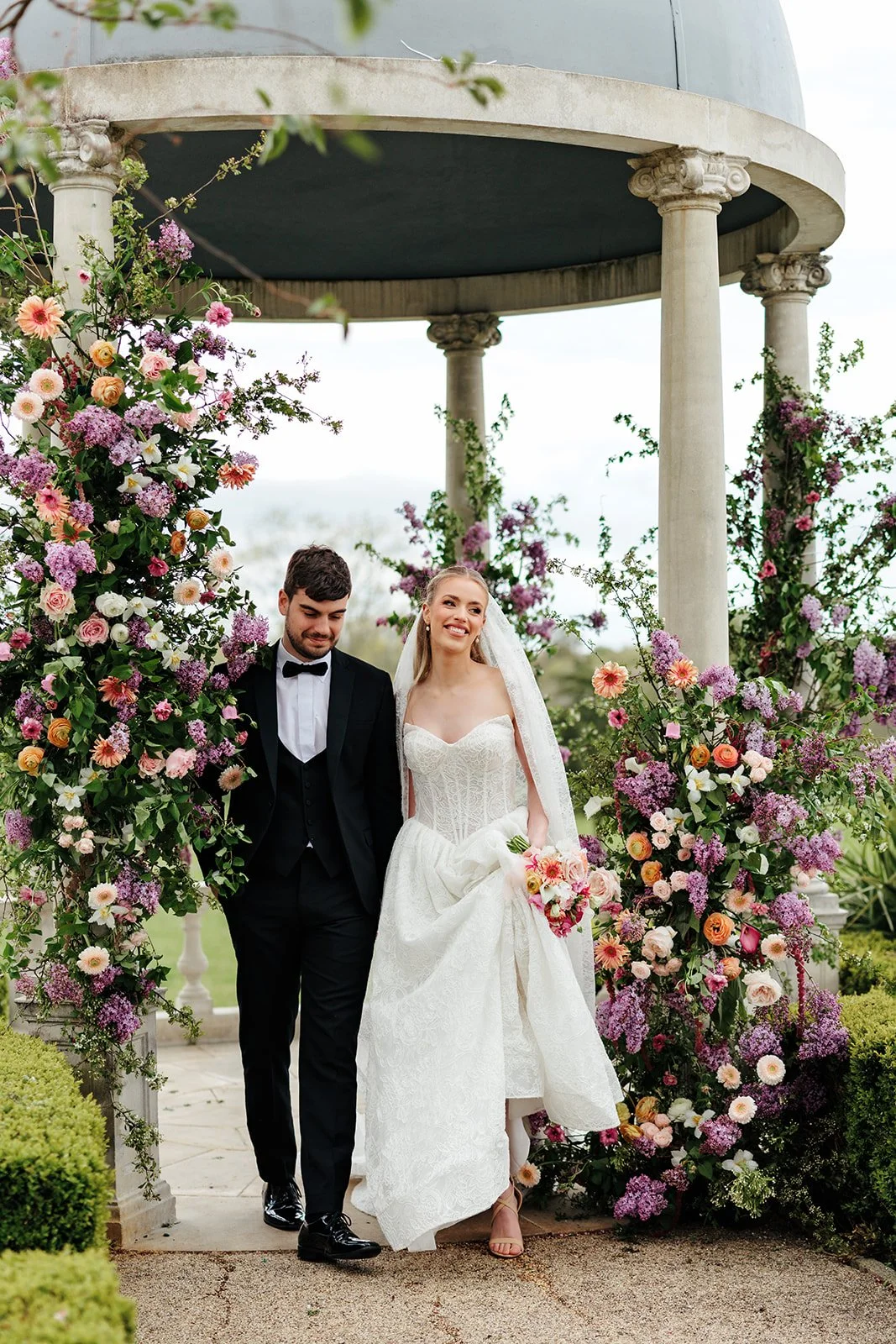 A bride and groom walking through a floral arch at an outdoor wedding ceremony, with the bride holding a bouquet and the groom in a black tuxedo.