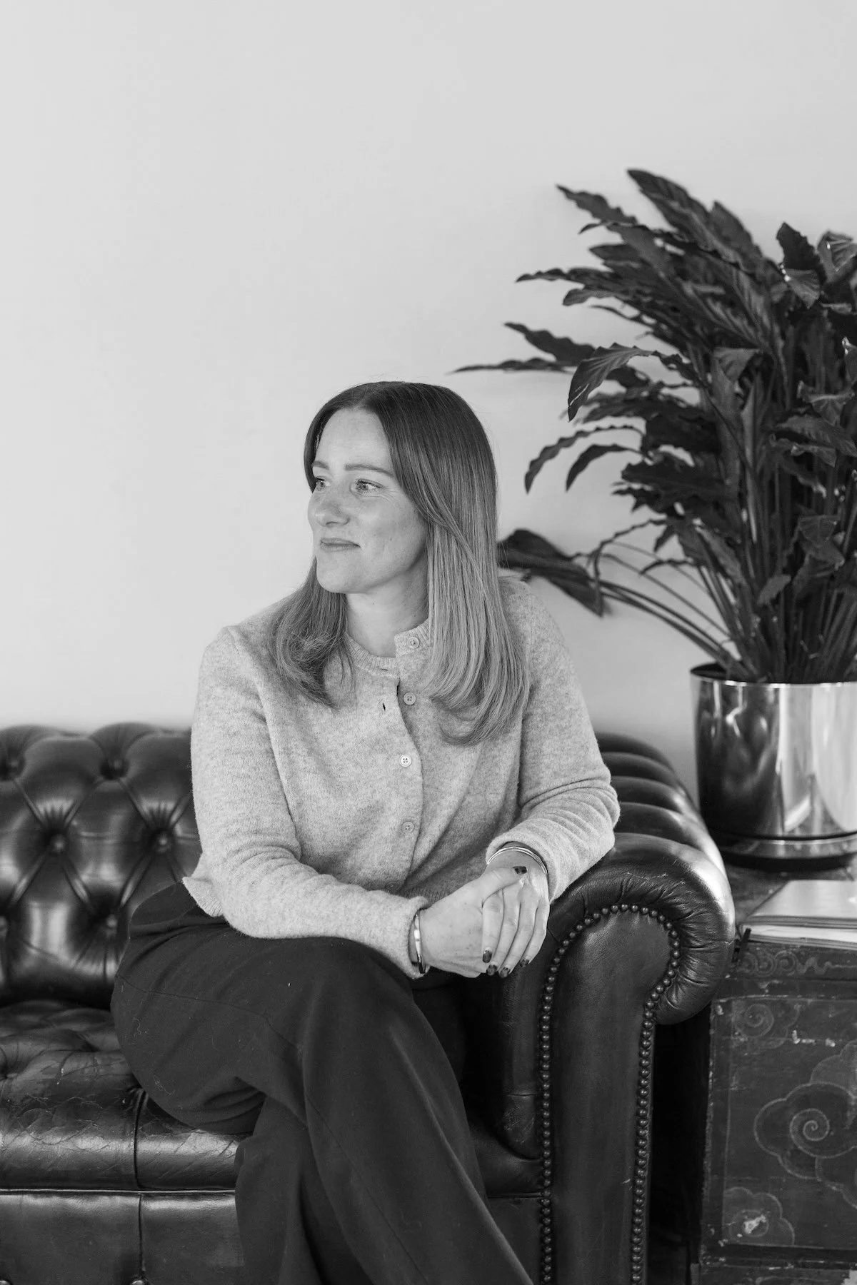 A woman with shoulder-length hair wearing a light-colored sweater and dark pants sitting on a tufted leather sofa next to a large potted plant.