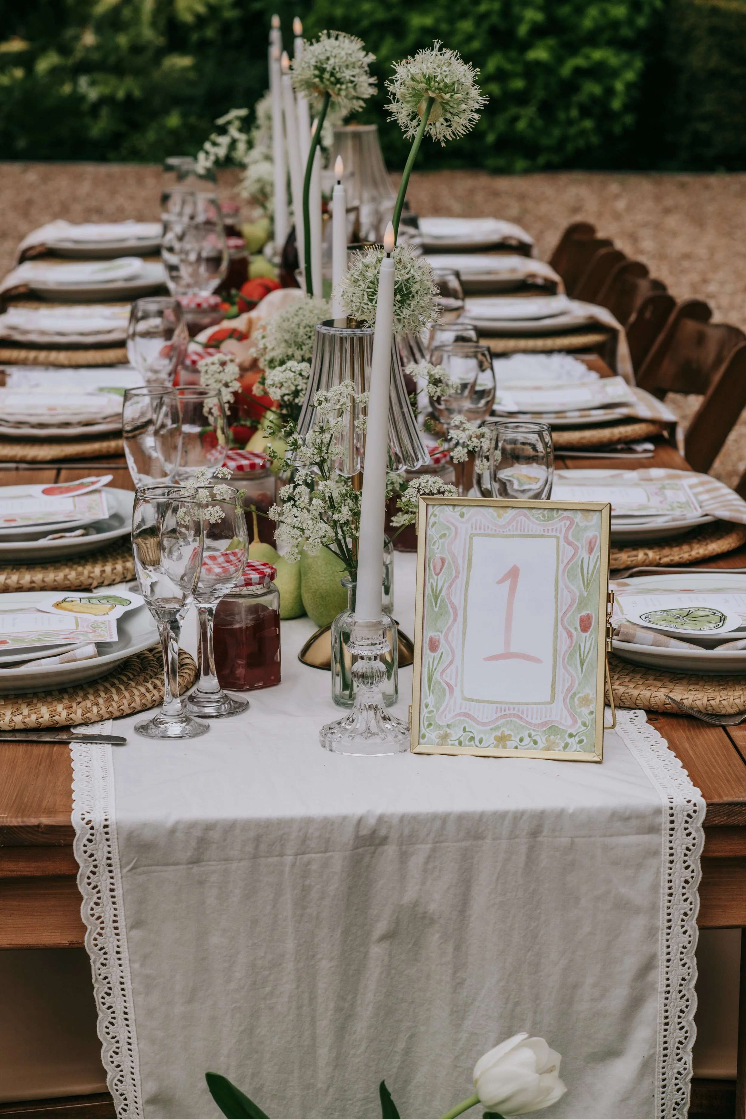 A beautifully set outdoor dining table decorated with white flowers, candles, and tableware, with a white lace tablecloth and a table number card reading '1'