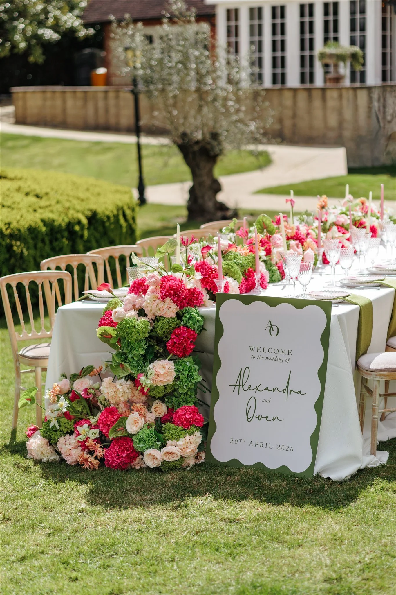 Outdoor wedding table decorated with pink and white flowers, a welcome sign with names Alexandra and Owen, and set with glasses, plates, and green napkins on a grassy lawn with a house and trees in the background.