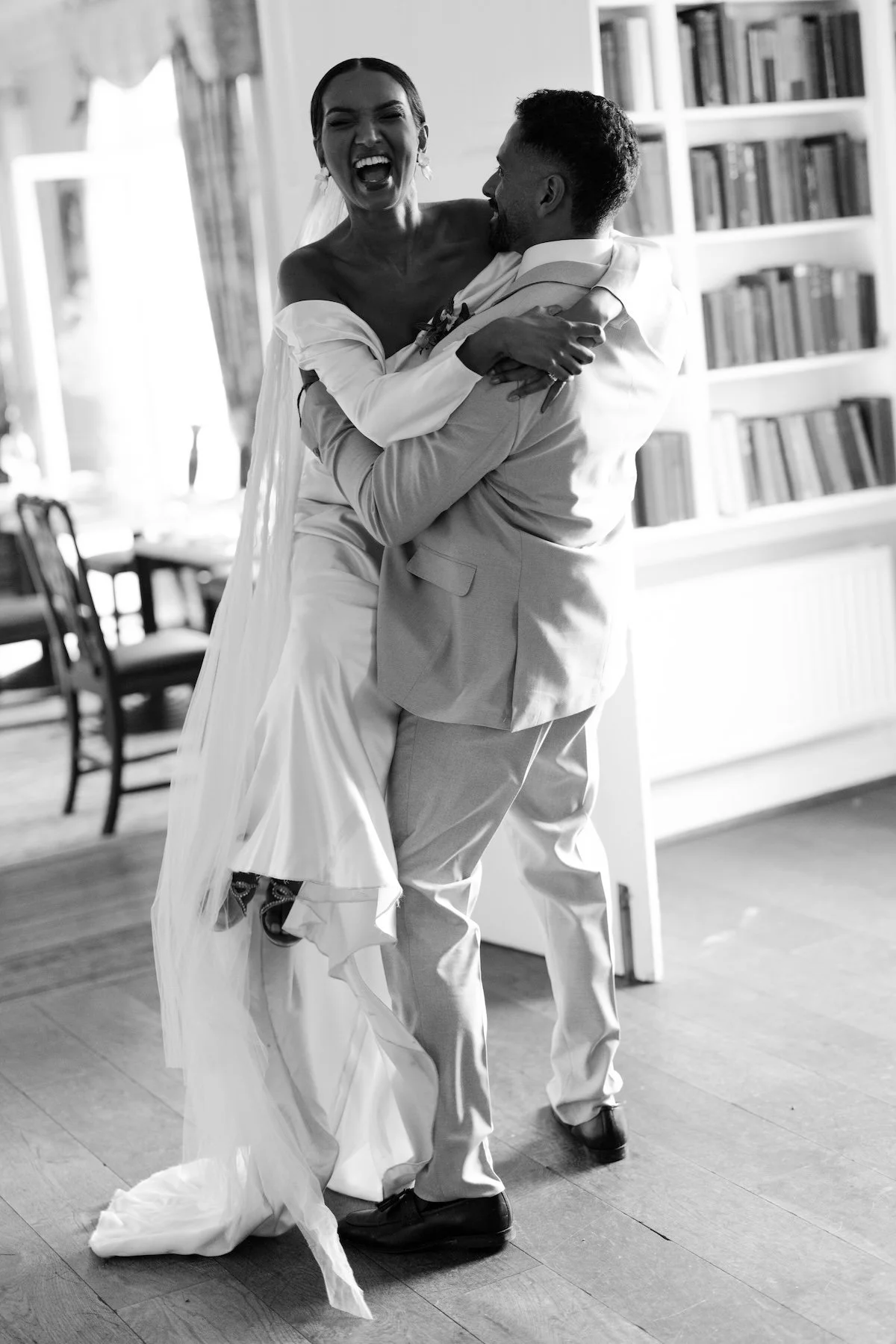 A joyful bride and groom dancing indoors, with the bride lifting her leg and smiling broadly, wedding attire, books on a shelf in the background.