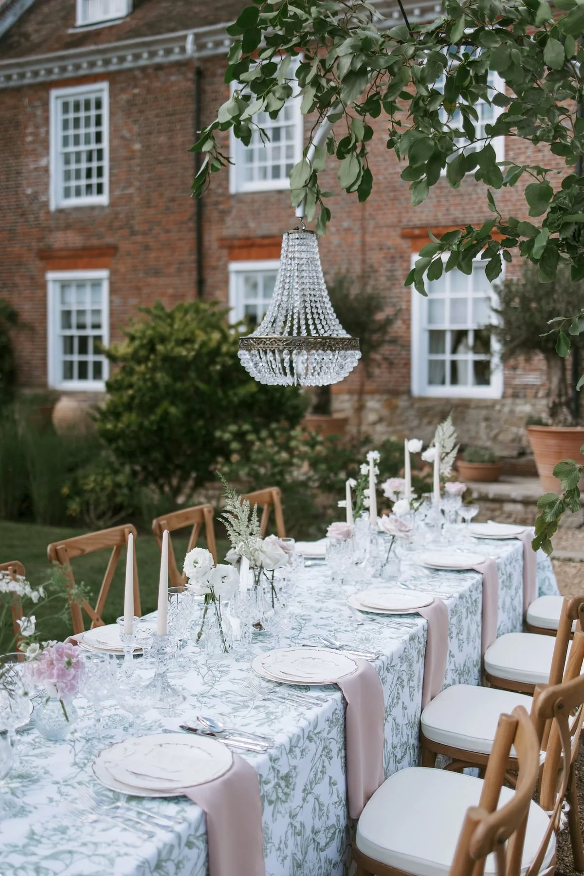 Outdoor dining table decorated with flowers and candles, featuring a chandelier hanging above in a garden setting with a brick house in the background.