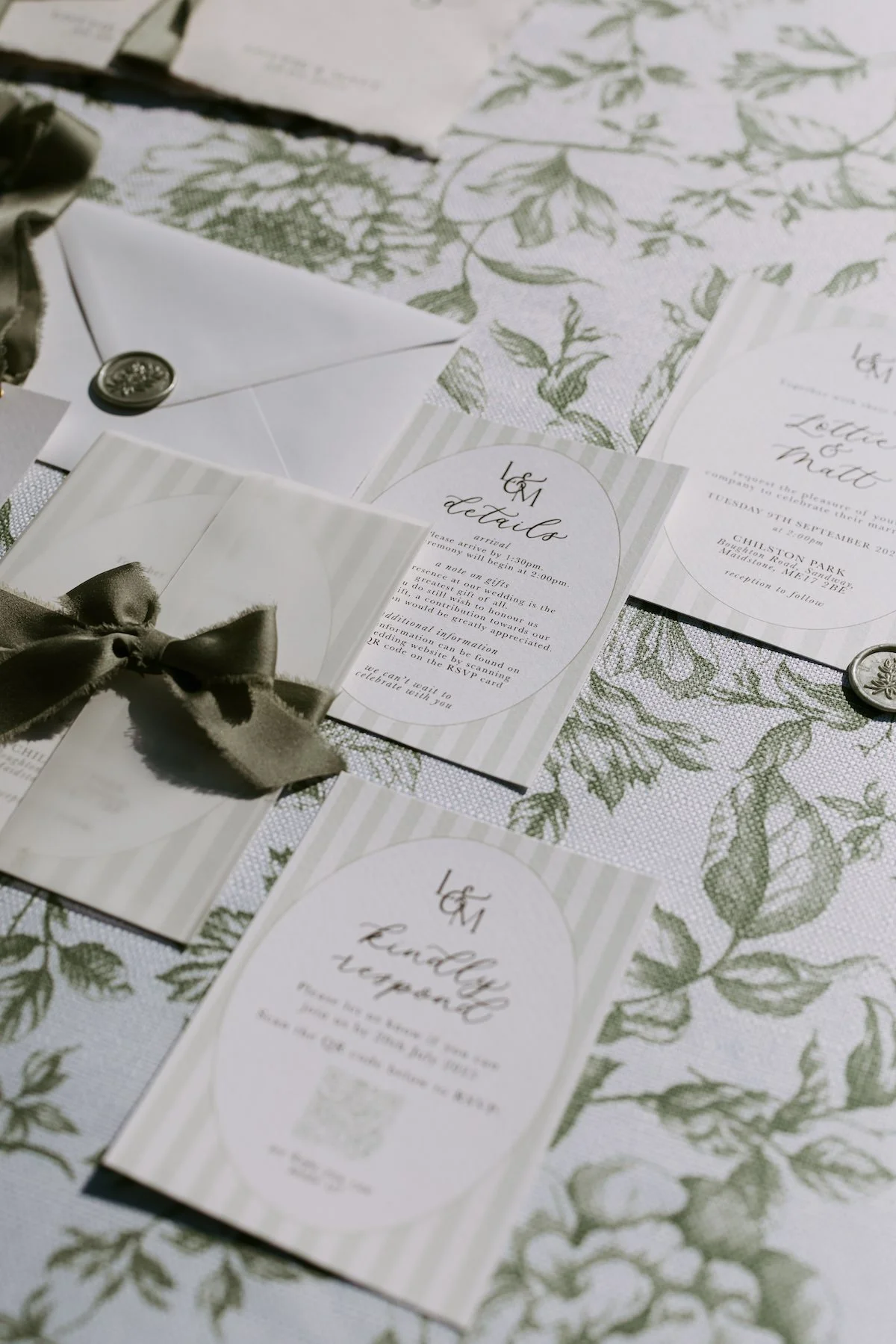 Wedding invitation suite on a floral tablecloth, includes details card, RSVP card, and envelopes with wax seals and bows.