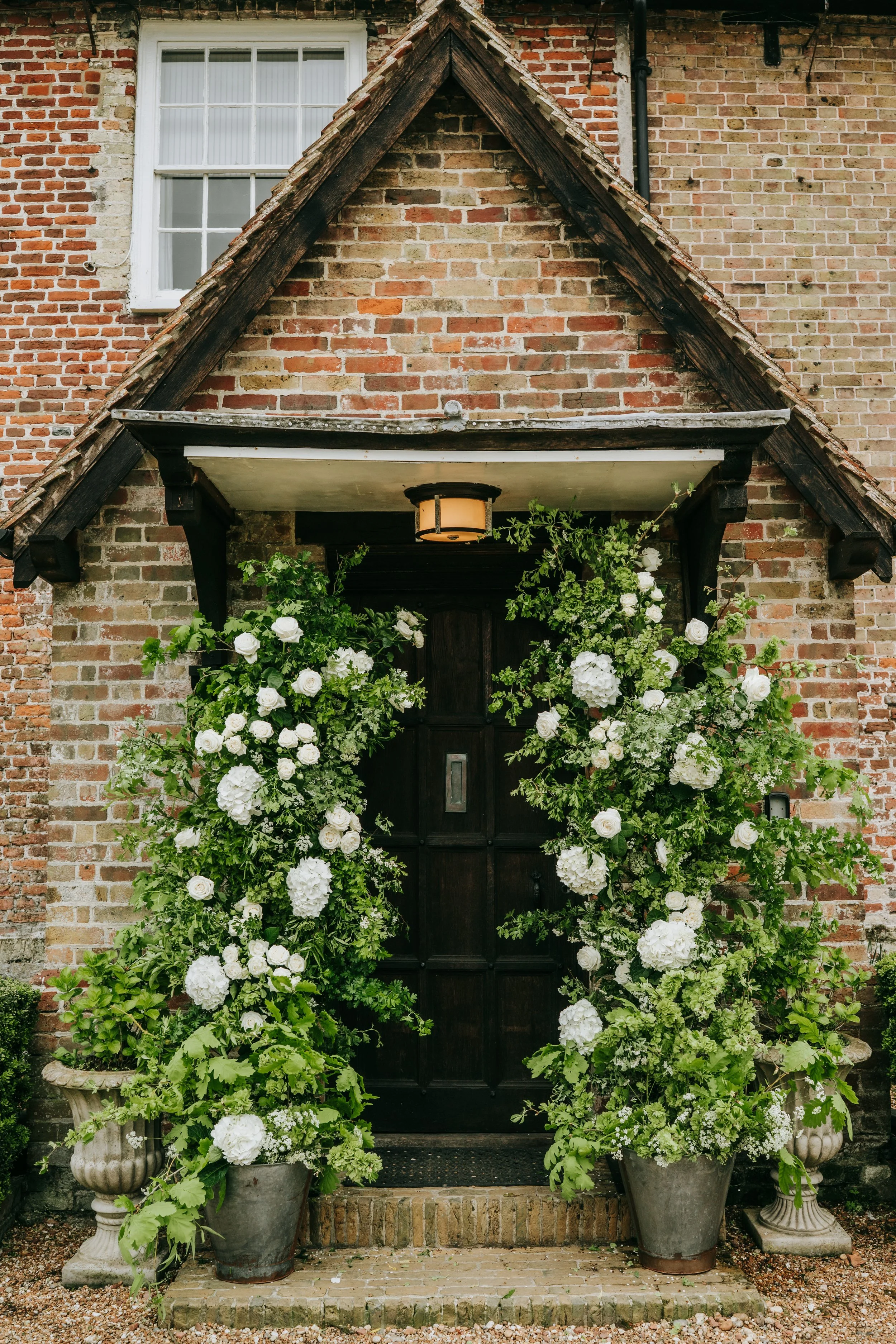 A black front door with floral arrangements surrounding it, placed in large decorative pots on either side of the entrance, with a brick wall, window, and porch light above.