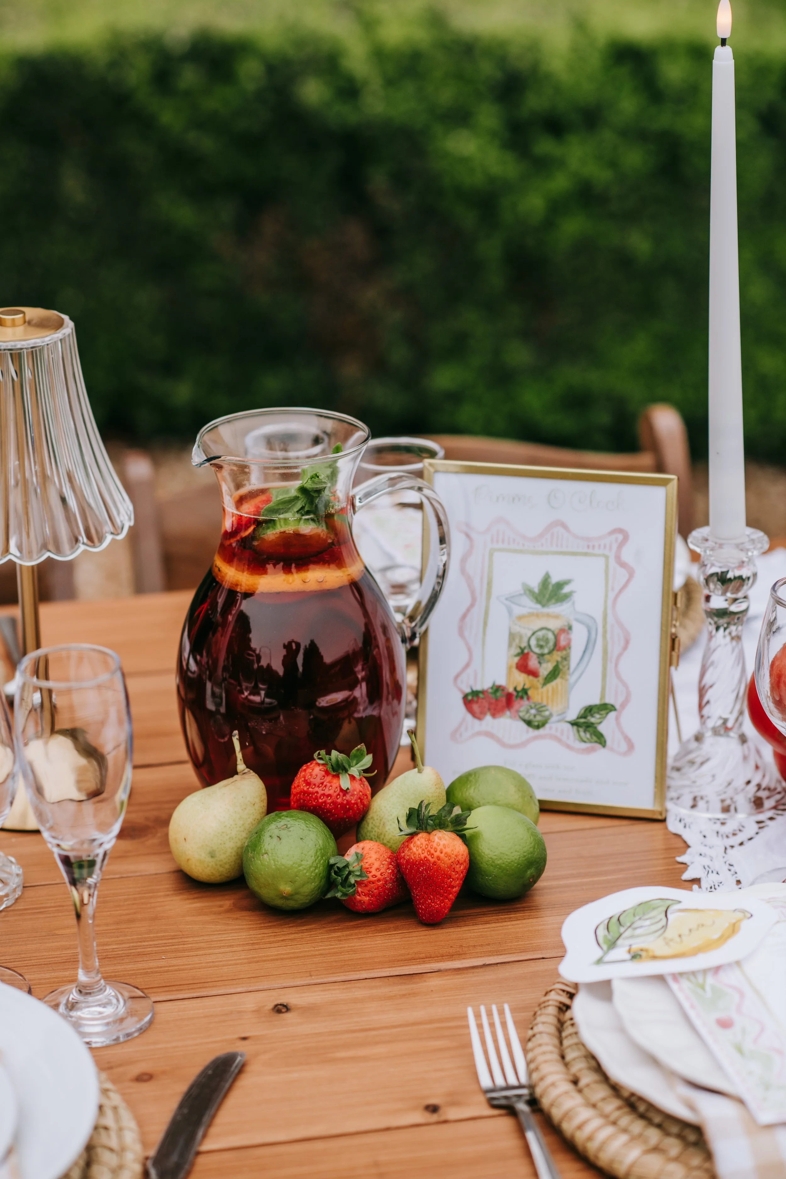 A table with a pitcher of red drink, fresh strawberries, pears, and limes, a glass, a candle, a menu, and tableware, set outdoors with a green background.