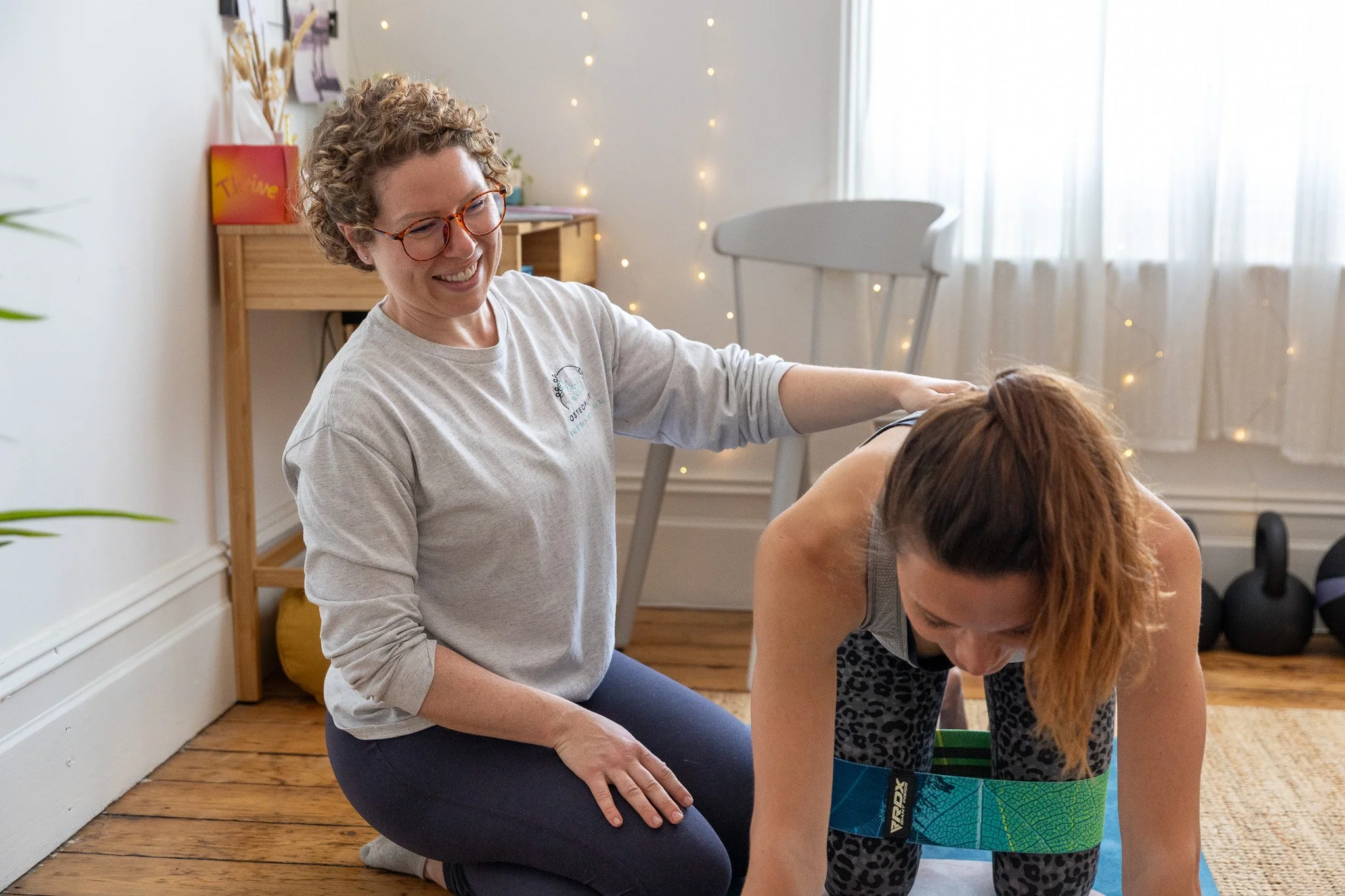A female Osteopath is assisting a woman in a workout position with a movement exercise on a yoga mat in a cosy room with fairy lights, a wooden shelf, kettlebells, and a window with white curtains.