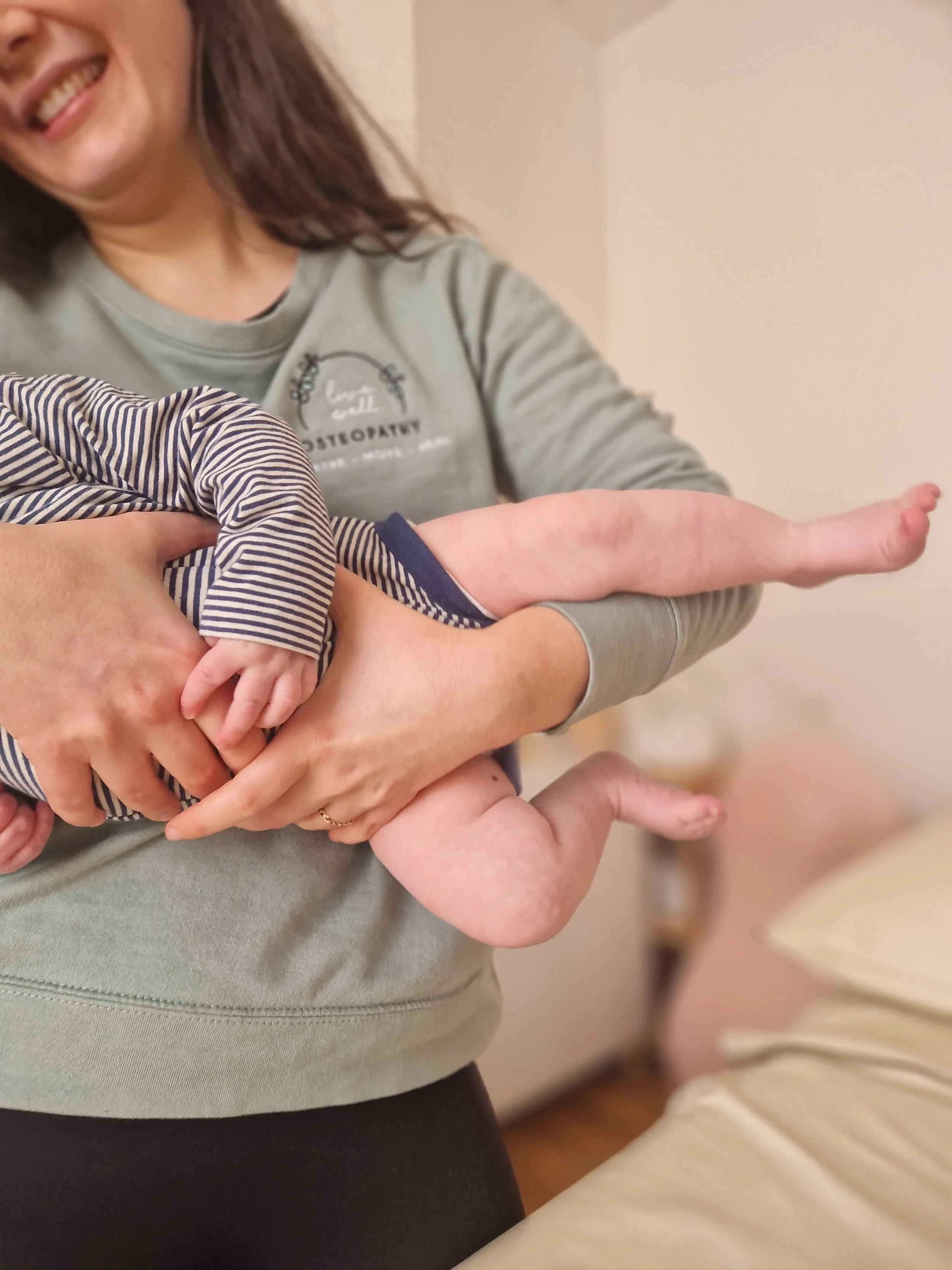 A female Osteopath holding a baby in her arms, with the baby's arm and leg extended outward. The woman is smiling, and the baby is wearing a striped outfit.