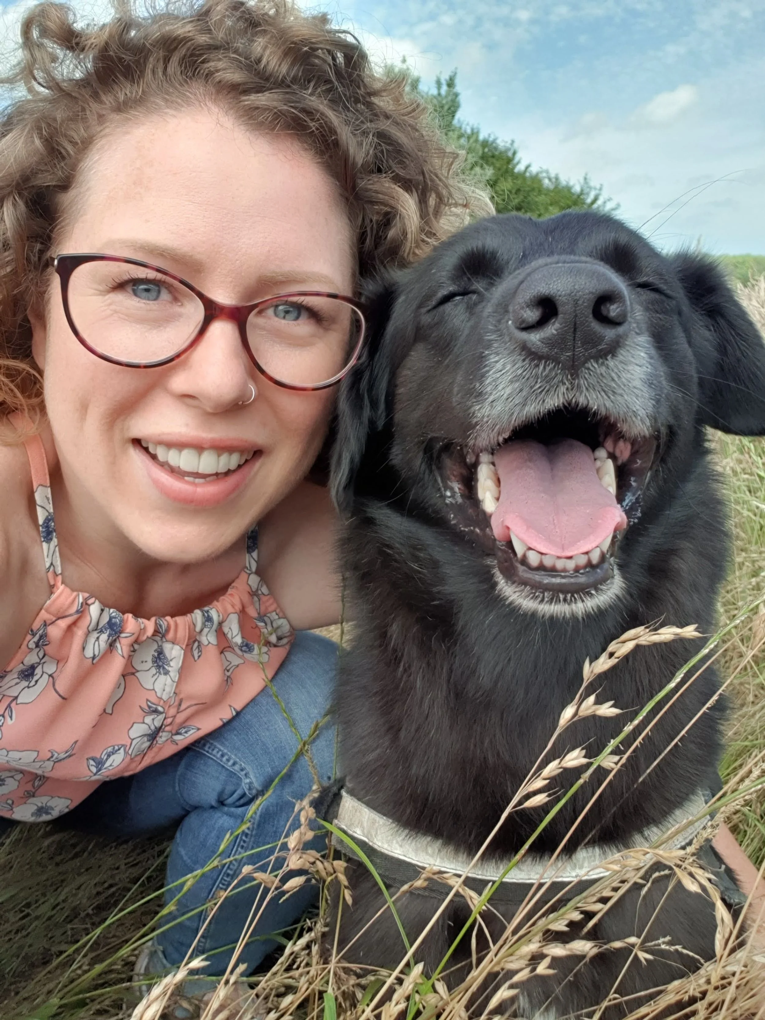 A woman with curly hair, glasses, and a nose ring smiling next to a black dog with its mouth open, in a grassy outdoor setting with a partly cloudy sky.