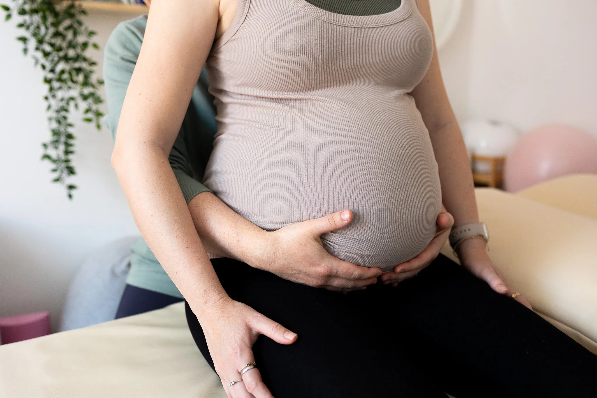 A pregnant woman in beige tank top sitting on a treatment couch with an Osteopath's hands supporting her belly in a cosy room with plants and soft décor.