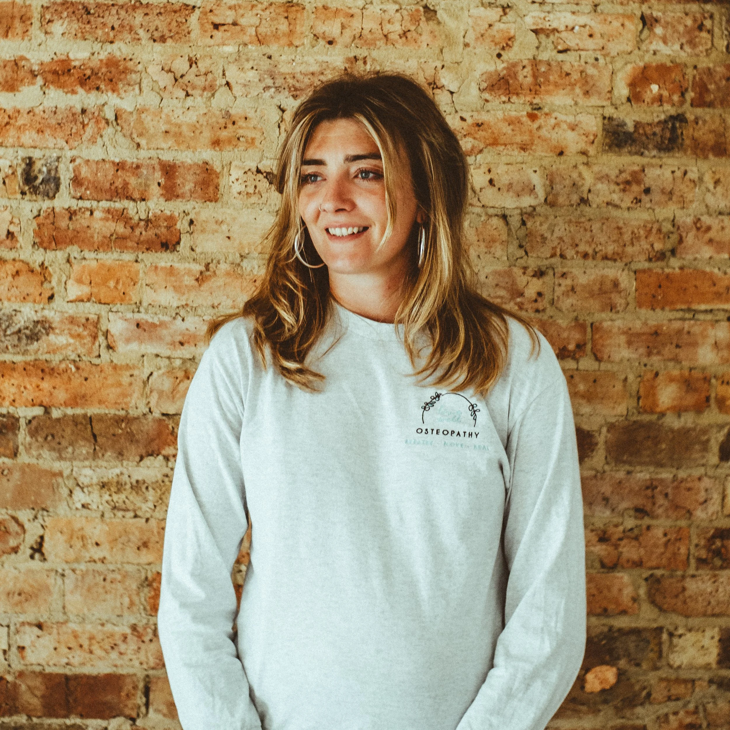 A young woman with light brown hair and hoop earrings standing in front of a brick wall, wearing a white long-sleeve shirt with a logo that reads 'Live Well Osteopathy'.