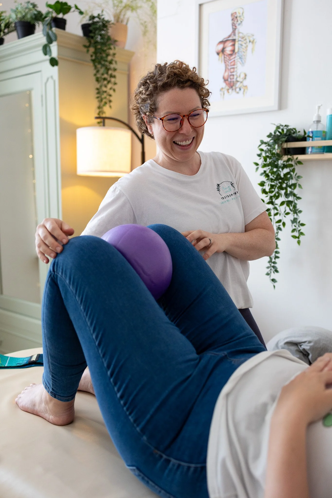 A cheerful female Osteopath smiling while performing a treatment using a purple massage ball positioned between a patient's knees in a well-lit room with plants and framed artwork.