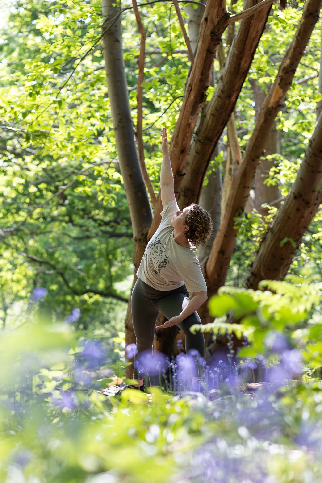 A woman practicing movement exercises outdoors among green trees and purple flowers in a forest.