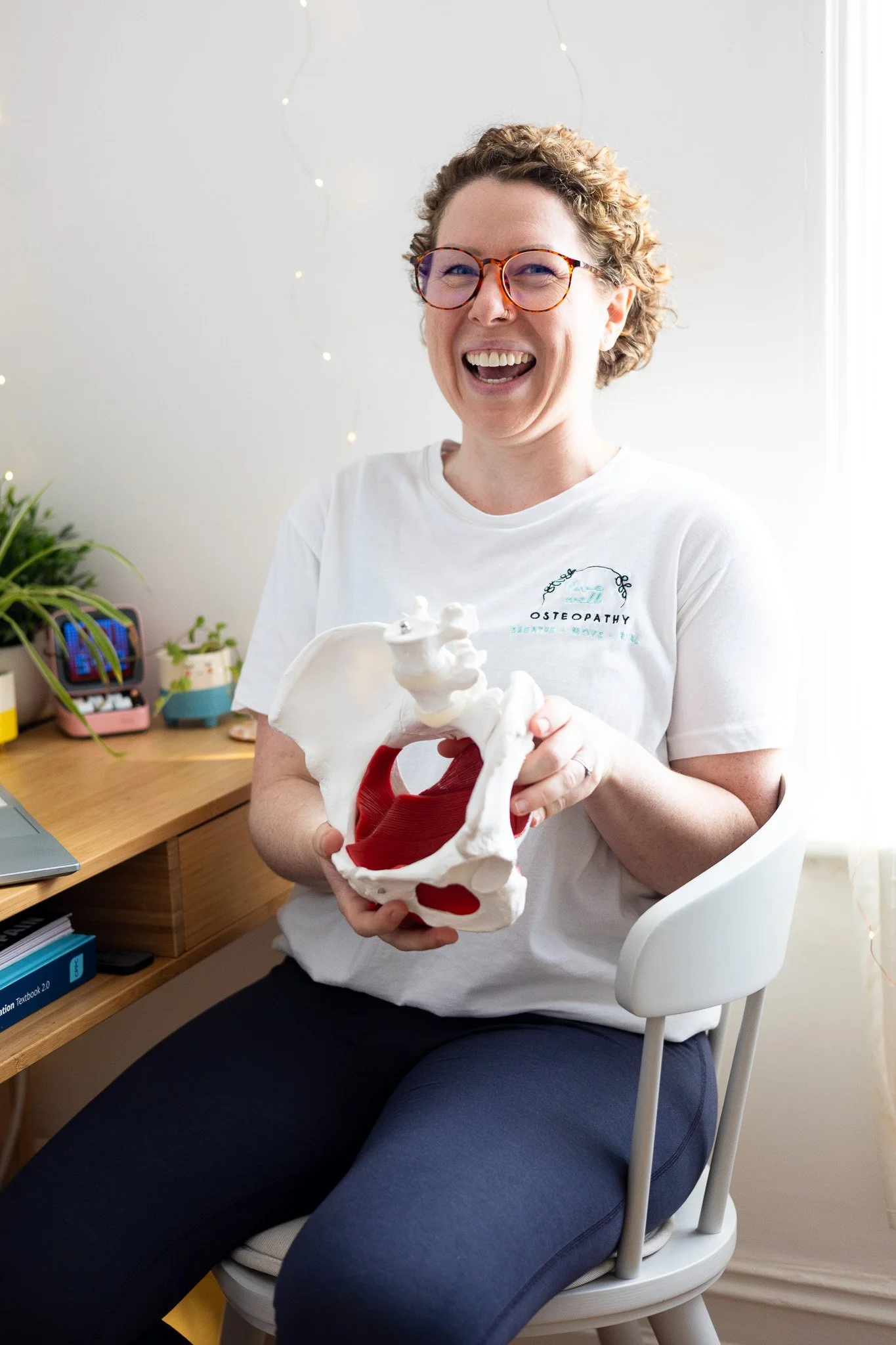 A female Osteopath with short curly hair, glasses, and a white t-shirt with the 'Live Well Osteopathy' logo sitting on a chair, smiling and holding a model of a pelvis with red muscles inside, in a room with a desk, plants, and fairy lights.