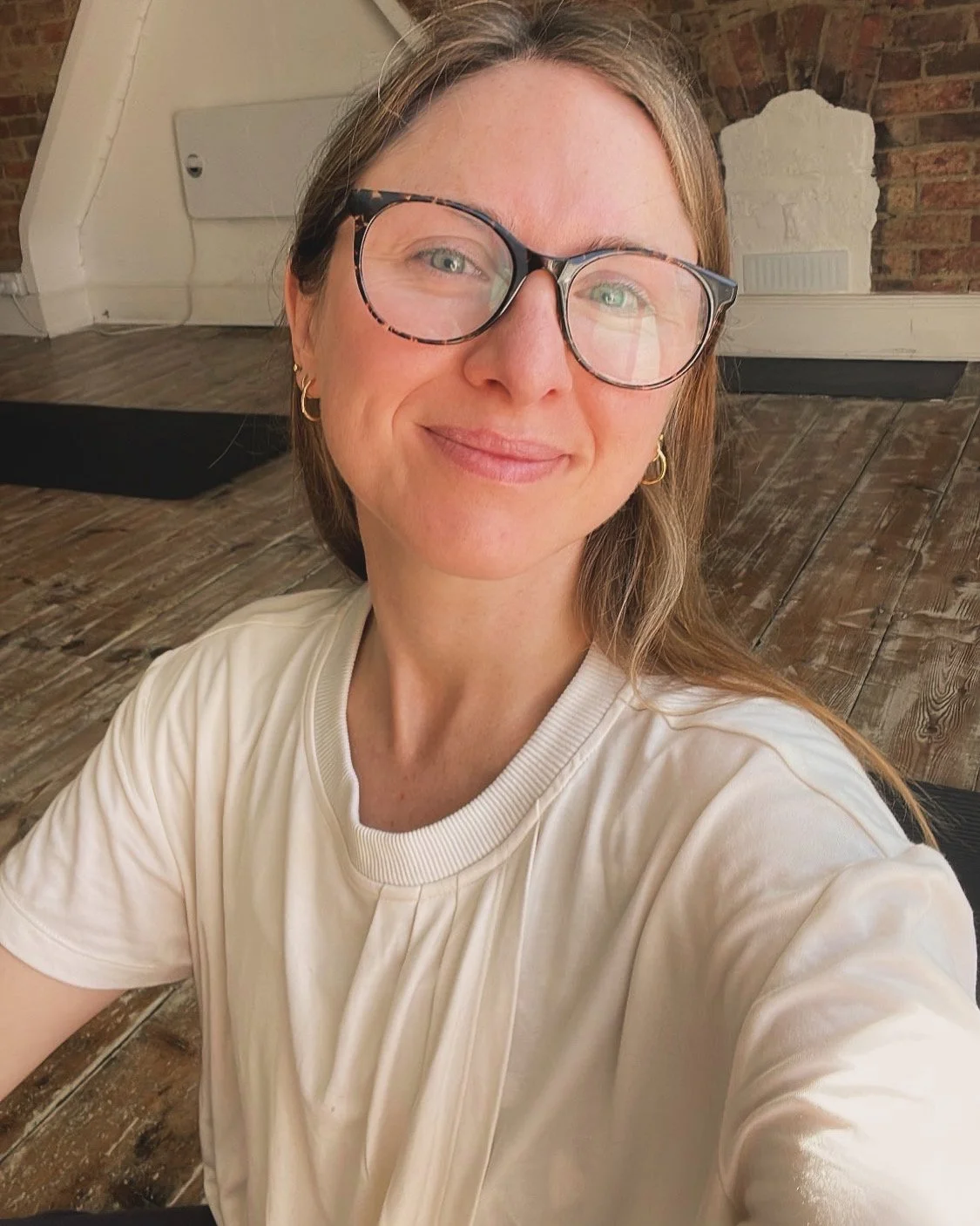A woman with glasses taking a selfie in a room with wooden floors and brick walls.
