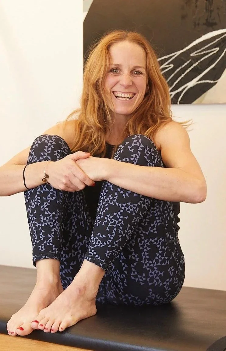 A woman in black workout clothes sits with her arms crossed around her knees. She smiles warmly at the camera.