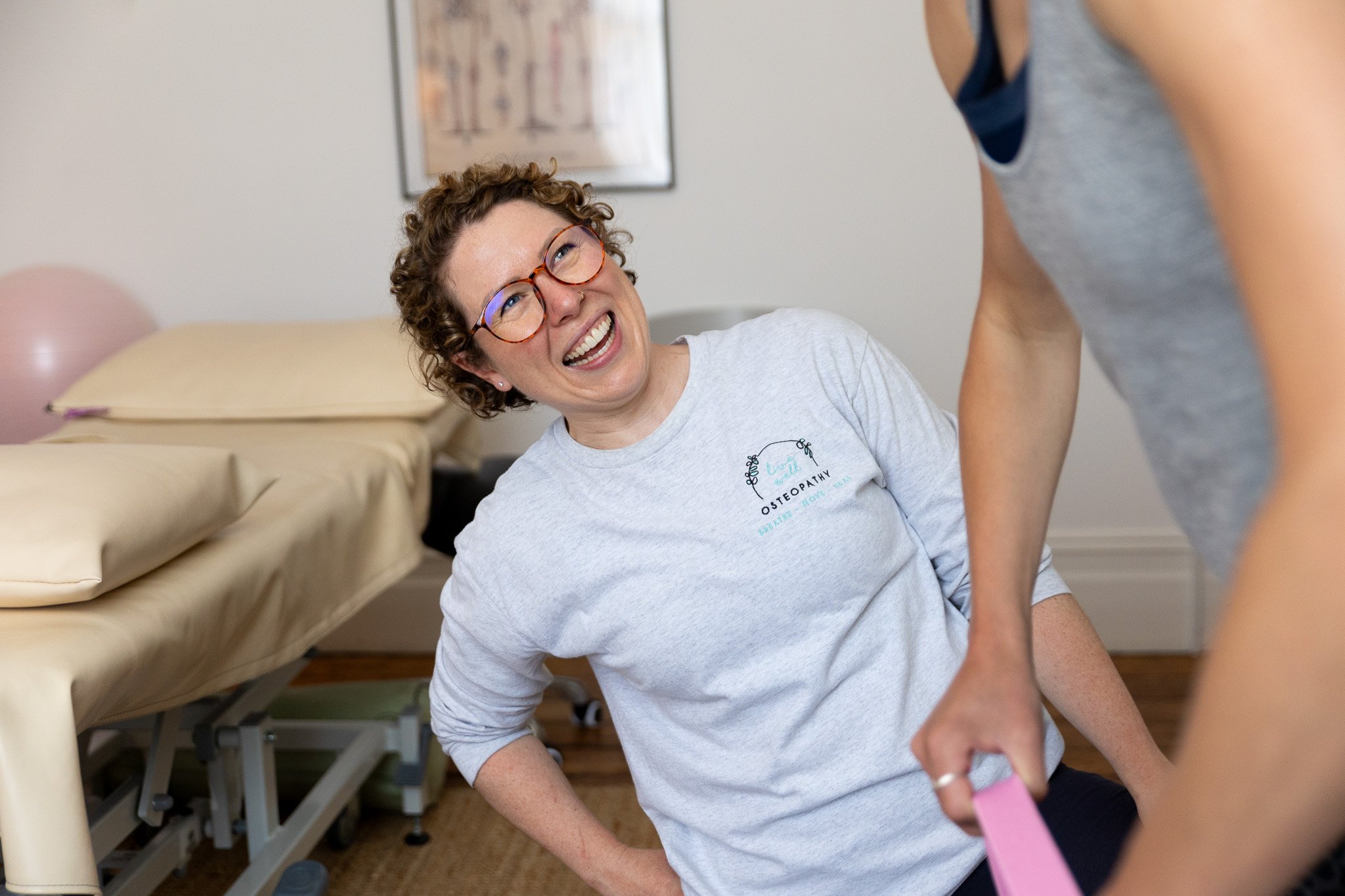 A female Osteopath with short curly hair, glasses, and a white t-shirt with the 'Live Well Osteopathy' logo smiles whilst demonstrating using a kettle-bell weight with a client.