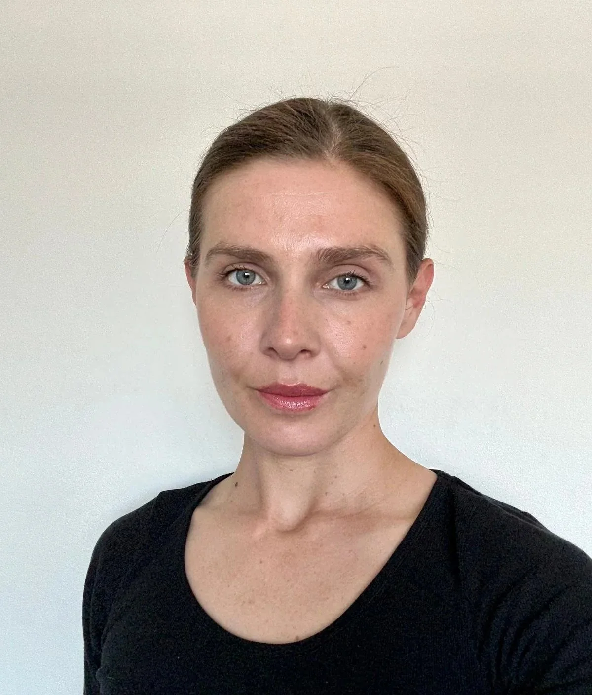 Close-up portrait of a woman with brown hair pulled back, blue eyes, wearing a black top, standing against a plain light-coloured background.