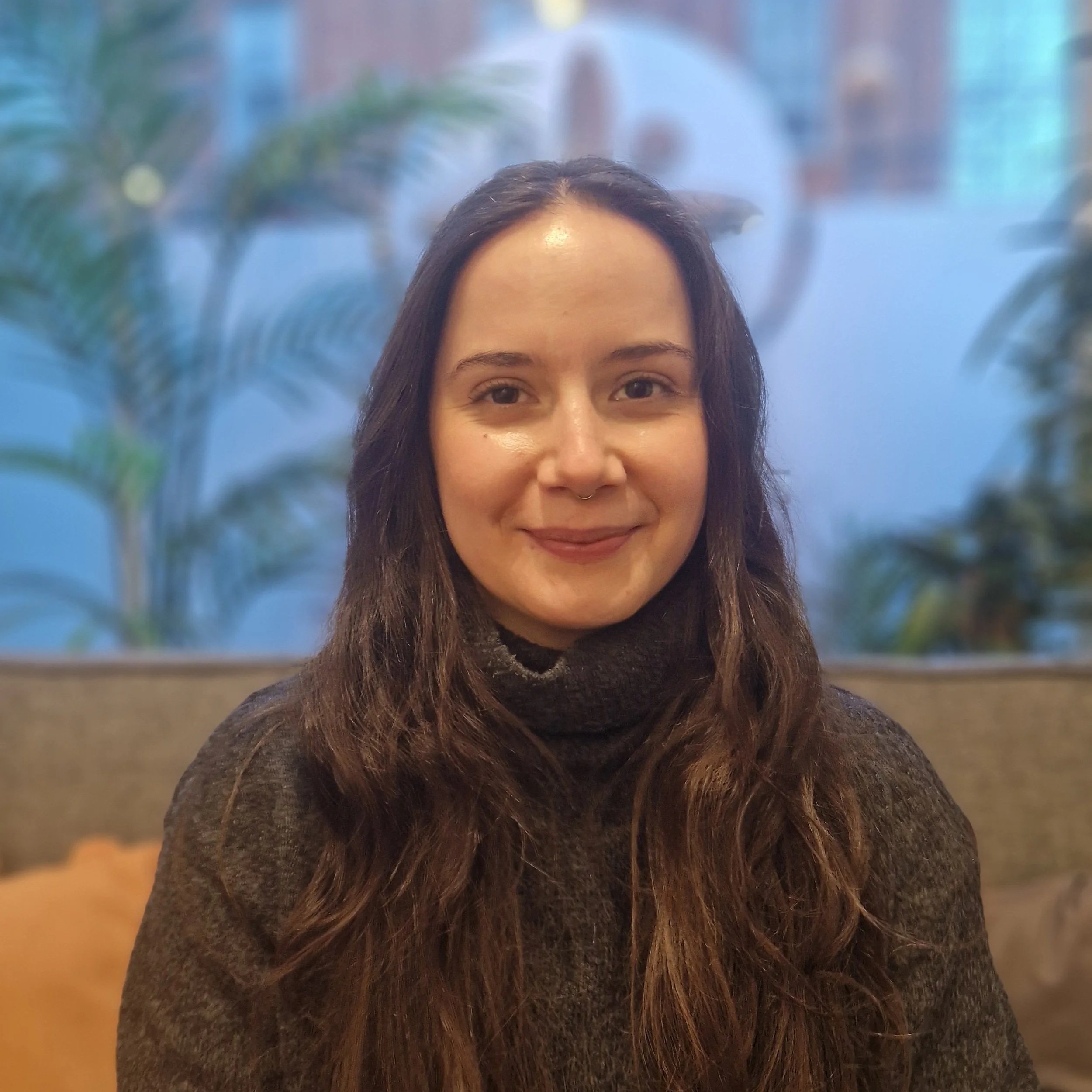 A woman with long brown hair and a black jumper smiling at the camera, with a blurred background of green plants and a large window displaying the Grace Hill logo.