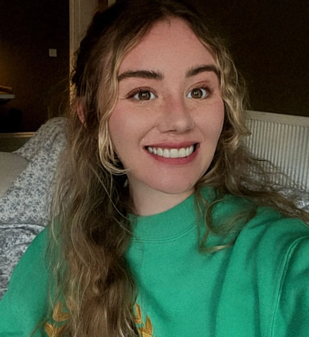 Emma smiles warmly at the camera. She has long hair and is wearing a green jumper.