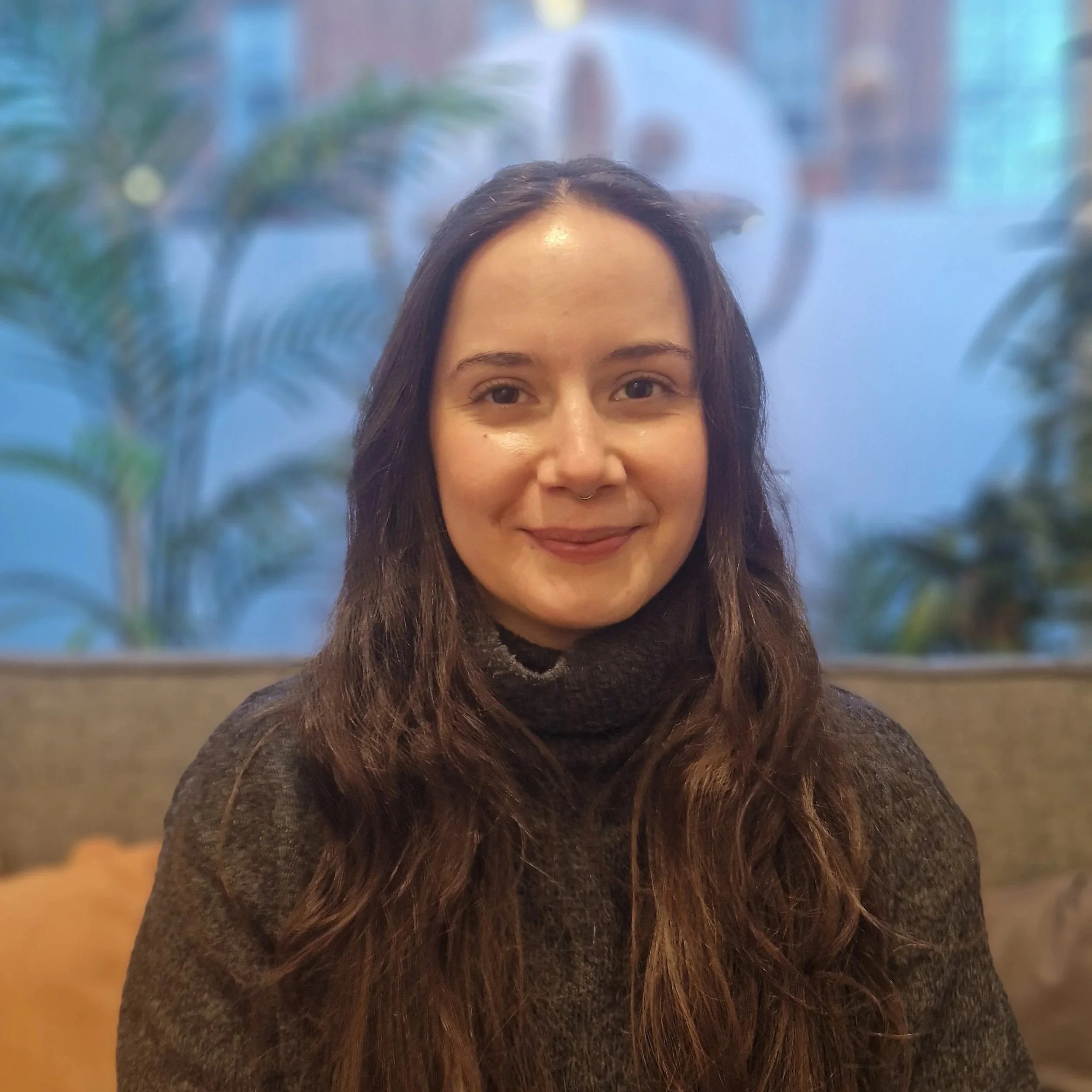A woman with long, wavy brown hair smiling in a cosy indoor setting with a large window displaying the Grace Hill Studios logo and plants in the background.