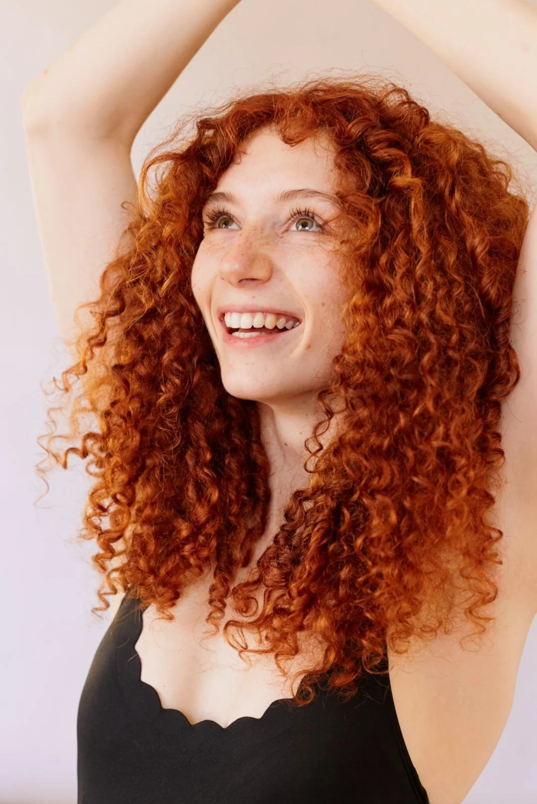 A woman with curly red hair smiling and looking up, with her arms raised above her head.
