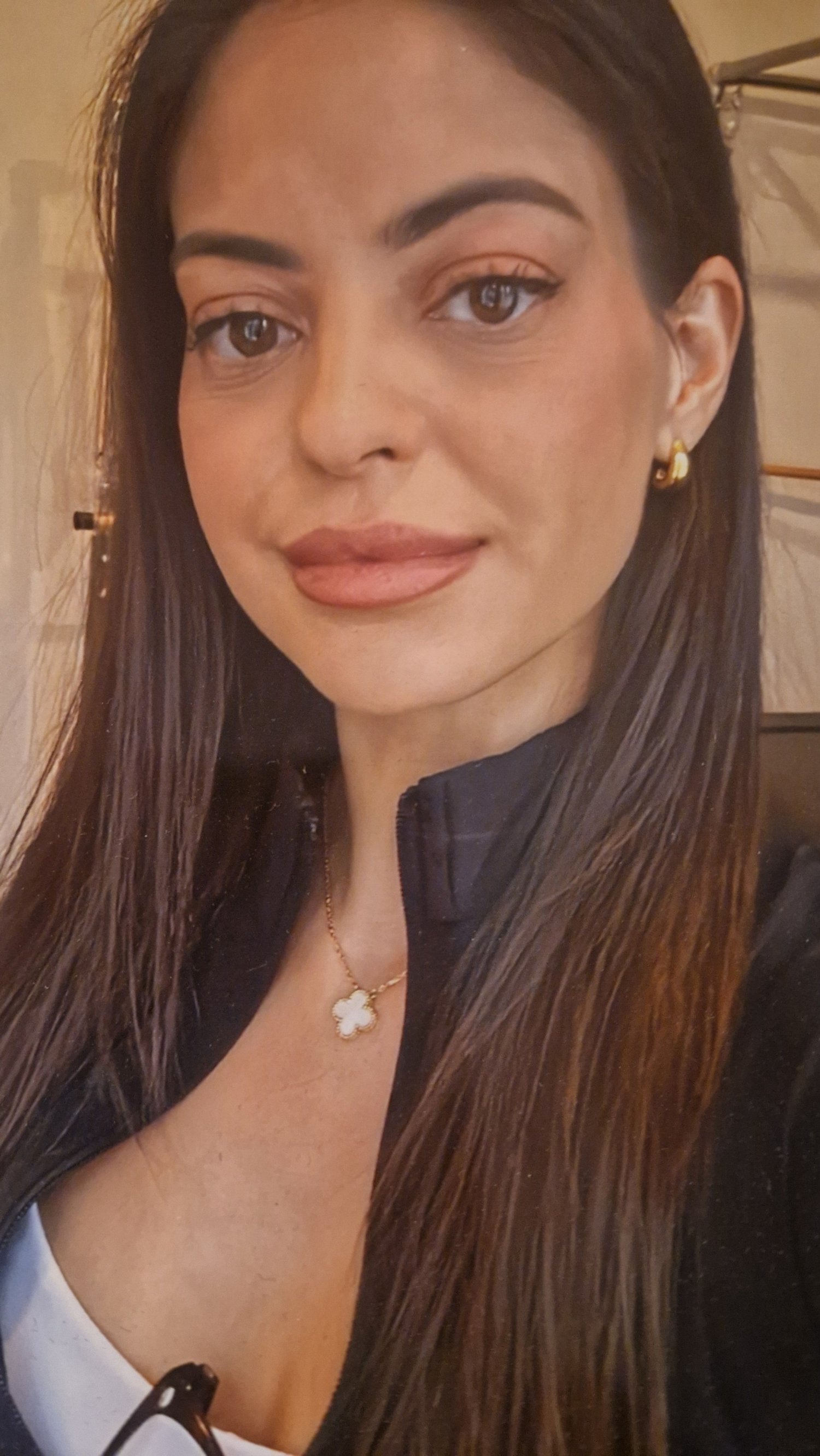 A woman with long dark hair, brown eyes, wearing gold earrings, a necklace with a clover pendant, and a black shirt.
