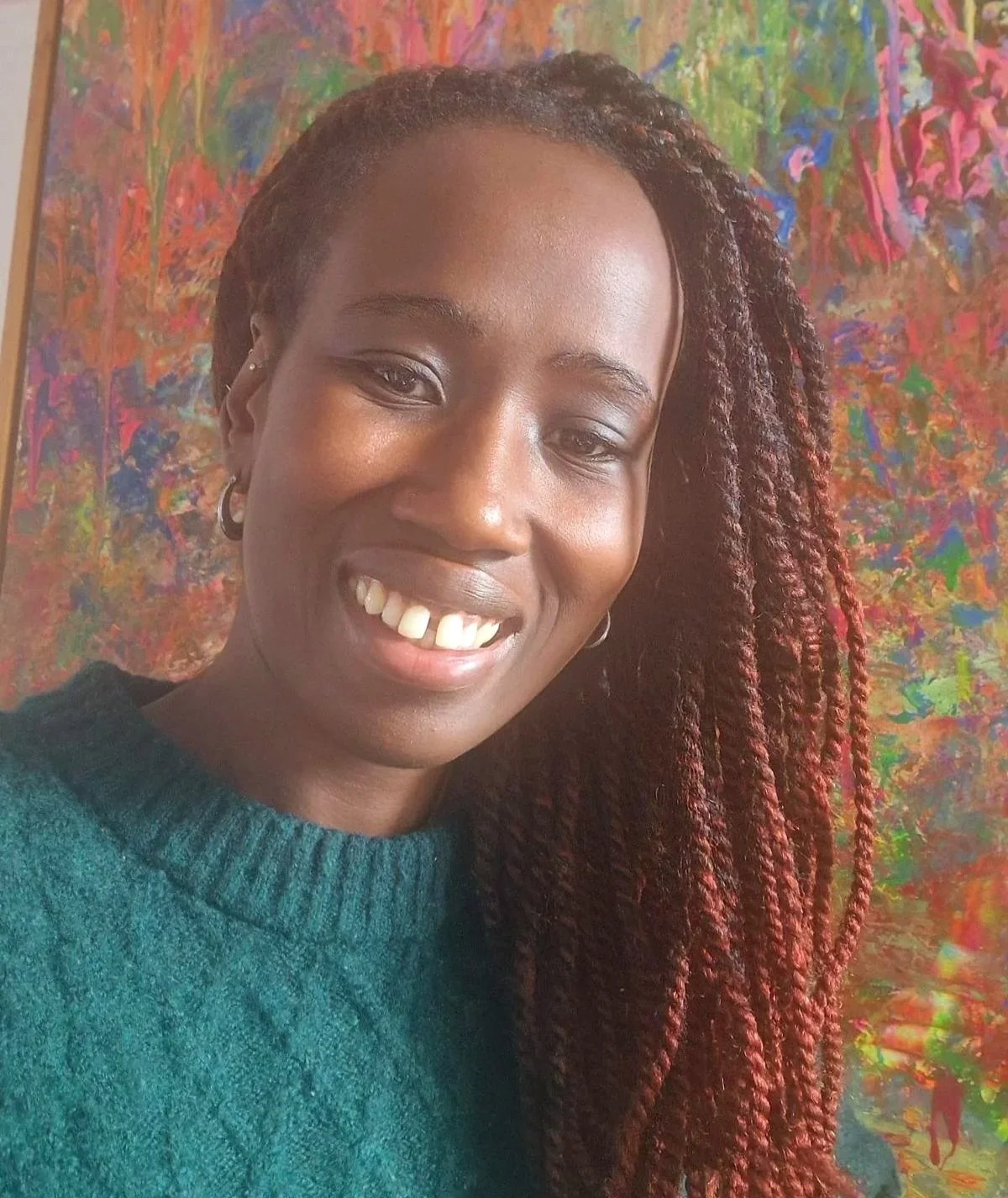 A woman with a big smile, wearing a green jumper, and having long red twisted braids, standing in front of an abstract multicoloured background.