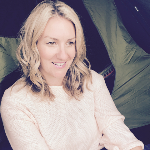 A smiling woman with wavy blonde hair, wearing a light-coloured jumper, in front of a green camping tent.